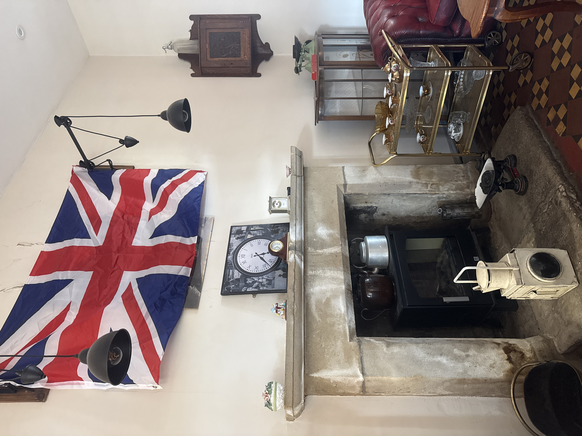 This is a photograph of the interior of a room, likely a living room or a study, decorated in a vintage or historical style, possibly in Lancaster, United Kingdom. Dominating the upper left of the frame is a large Union Jack flag, draped across the wall and affixed with what appears to be a pulley system for adjustment. Two black industrial-style pendant lights hang from the ceiling, one directly over the flag and another on an articulated arm extending from the wall. Below the flag, a stone fireplace mantelpiece is adorned with several objects. On the left, a small, ornate ceramic dish with a floral motif sits on the mantel. To its right, a small, colorful figurine of what appears to be a carousel horse is placed. Further to the right, a framed picture contains a large clock face with Roman numerals, surrounded by smaller black and white images, suggesting a collage or montage. A smaller, rectangular clock with a gold frame sits to the right of the framed picture. Within the fireplace opening, a black wood-burning stove is visible, with a dark brown teapot on the left and a silver kettle on the right, both positioned on top of the stove. In front of the stove, to the right, a white, antique-style lantern with glass panels and a handle is placed on the hearth. To the left of the fireplace, a black metal bucket or coal scuttle is positioned. To the right of the fireplace, a tall, glass-fronted wooden cabinet stands in the corner, containing several items. On top of the cabinet, a vintage green mechanical scale with two platforms is visible, along with a red box. A wooden corner cabinet with decorative carvings is mounted on the wall above the glass cabinet. In the foreground, a gold-colored three-tiered tea trolley is laden with teacups, saucers, a teapot, a sugar bowl, and a milk jug on the top shelf. The middle and bottom shelves also hold glassware and bowls. To the far right, a portion of a deep red leather sofa is visible, and a small wooden table with a reddish-brown top is partially in frame. The floor is tiled with a pattern of alternating brown and cream squares. The overall impression is one of a room that has been curated to evoke a specific historical period, possibly a tea room or a collector's space. There are no people