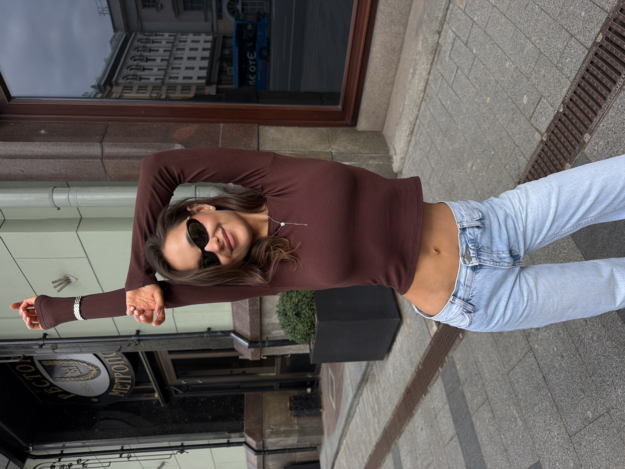 A young woman is captured in a full-body portrait on a city sidewalk, posing with her arms raised and bent behind her head. She faces the viewer with a faint smile, wearing dark sunglasses, a close-fitting dark brown long-sleeved crop top that exposes her midriff, and light blue high-waisted jeans. A delicate silver necklace with a small pendant adorns her neck, and a silver watch is visible on her left wrist.
The setting is an urban environment, likely in Moscow, Russia, as indicated by several visual cues. The woman stands on a paved sidewalk composed of grey and lighter grey square and rectangular tiles. A metal drainage grate with the partial text "STANDARD PARK" is embedded in the sidewalk to her right. Behind her is a building with large, dark-framed windows that reflect the opposite side of the street and the sky.
Reflected in the window are architectural details of a grand building, an overcast sky, and a blue bus with "МЕОТЕ" (partially visible, likely part of "МЕТРОПОЛИТЕН," meaning Metropolitan/Metro) in Cyrillic. Crucially, a large sign reflecting in the window clearly displays "РЕСТОРАН МЕТРОПОЛЬ" (Restaurant Metropol), a famous establishment in Moscow, confirming the location. Below the window, there's a dark rectangular planter holding a green, manicured shrub. The time of day appears to be daytime, possibly overcast, given the subdued light and the reflected sky. The overall mood is casual and stylish.