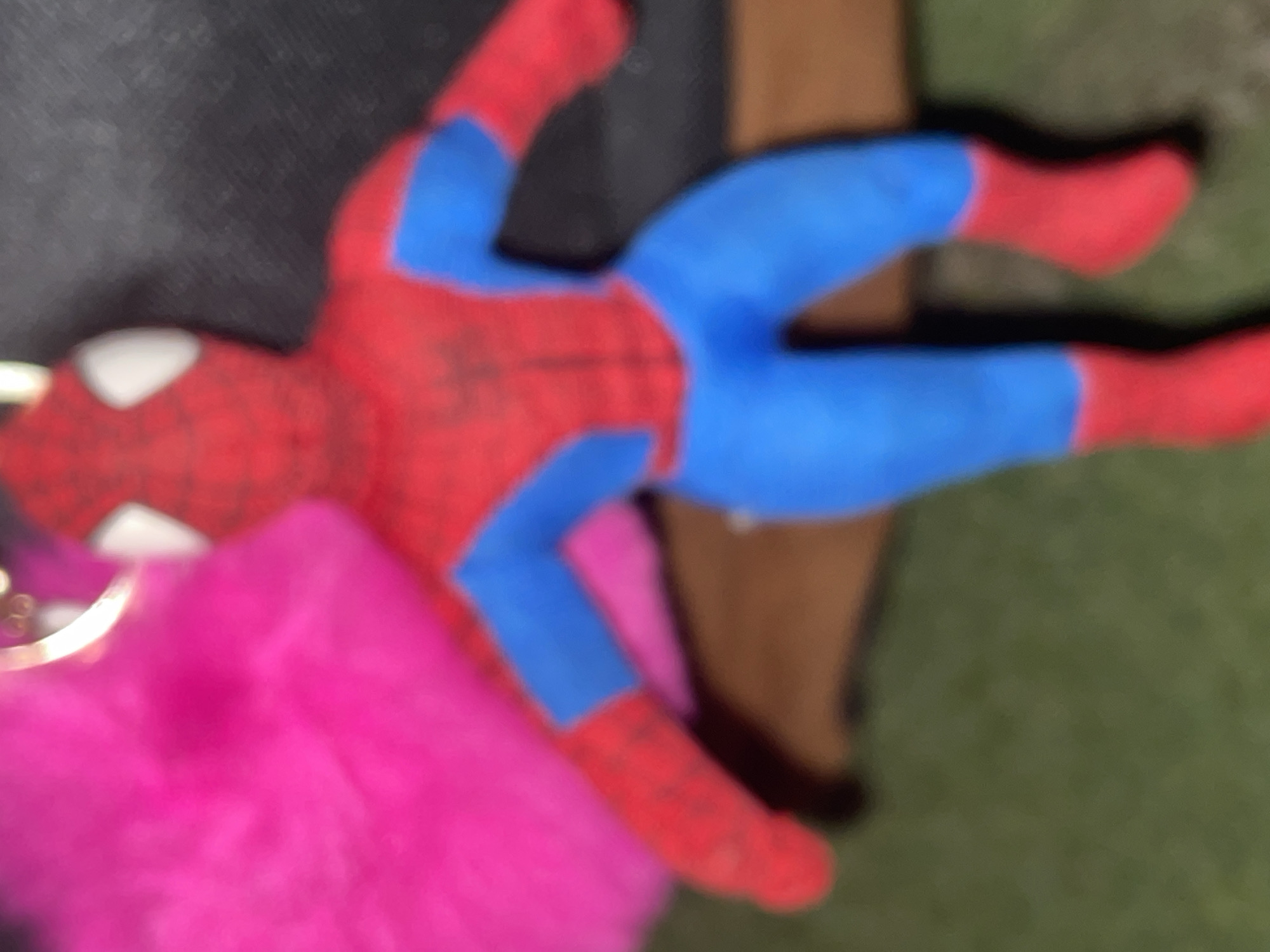 In the city of Santa Ana, Venezuela, a blurry close-up depicts a soft Spider-Man plush toy in a seated position. The toy features a red body with blue limbs, red gloved hands and boots, and white eye outlines on its masked face, with a faint web pattern visible on its chest. To the upper left, a vibrant, out-of-focus pink fluffy or furry object is partially visible, attached by a golden metallic ring, suggesting a keychain or accessory. The background includes a dark, indistinct surface behind the toy, transitioning to a blurry brown surface beneath it, and an out-of-focus green ground cover at the very bottom, resembling grass.