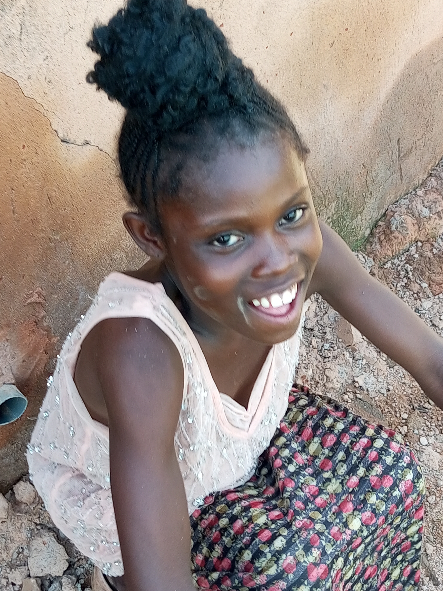 A young Black girl with her hair pulled up in a bun is smiling broadly. She is wearing a light pink sleeveless top adorned with sequins and a floral patterned skirt. She is seated outdoors with a textured, light-colored wall behind her and rough, rocky ground beneath her. The scene appears to be in the city of Tempure, Nigeria.