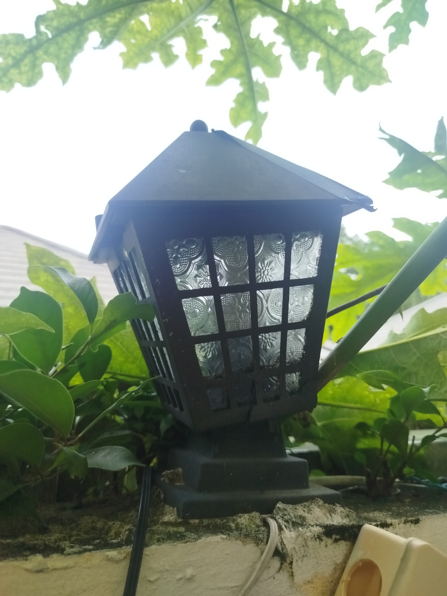 In Baan Lamai, Thailand, a black, lantern-style outdoor light fixture is mounted on a textured, light-colored wall. The fixture has decorative, patterned glass panels within a grid structure. Green foliage, likely from a tree with large, lobed leaves, frames the top and sides of the scene, with some leaves appearing to be backlit by the sky. The overall environment suggests an outdoor setting in a tropical or subtropical region, potentially a garden or courtyard, with the fixture positioned against a wall that exhibits some signs of weathering and moss growth. A black wire is visible near the base of the fixture.