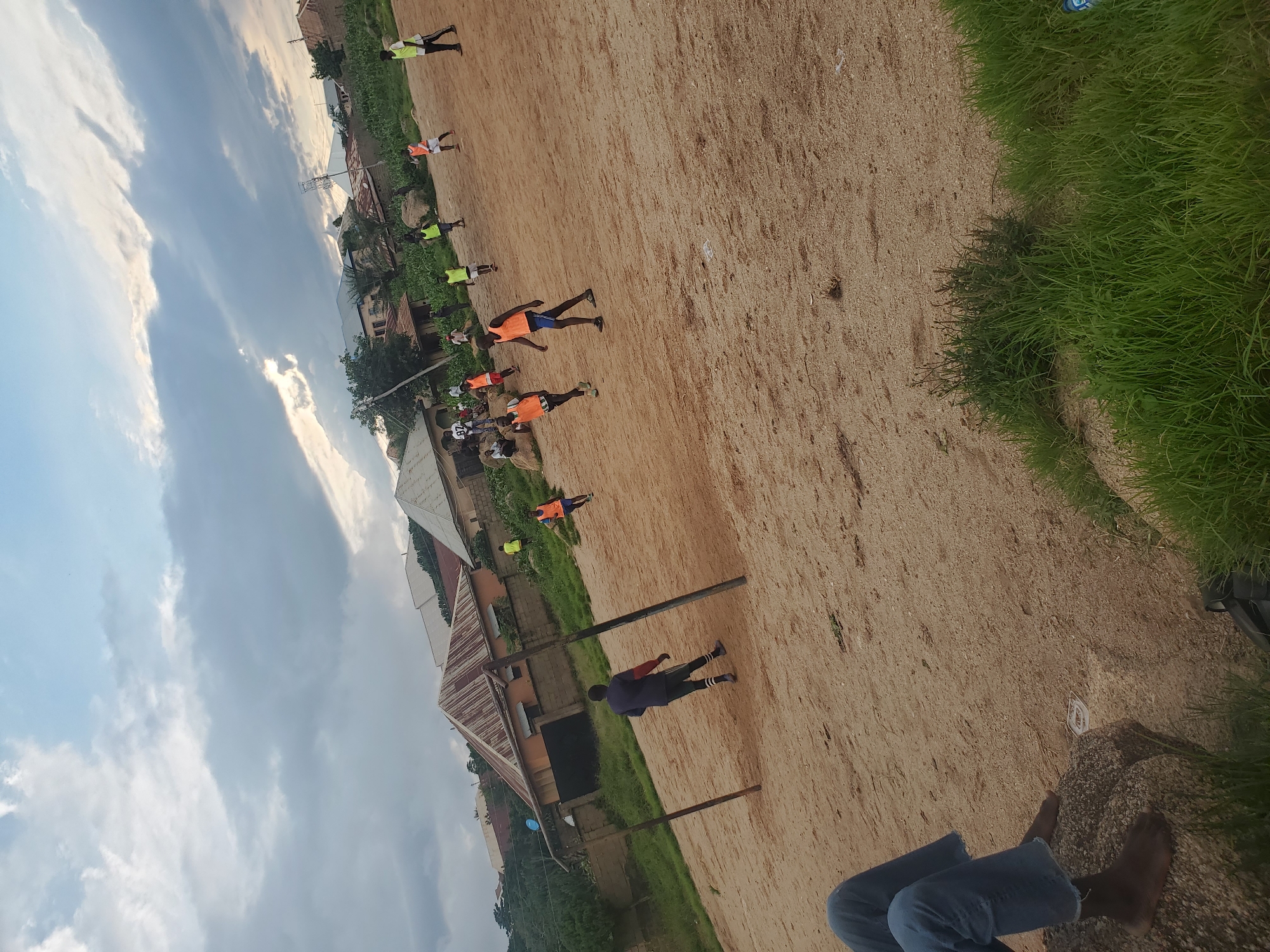 An aerial outdoor daytime shot shows a group of young people playing soccer on a dirt field. A person in a blue shirt and dark shorts stands in the foreground on the left, facing away from the camera. They are barefoot and sitting on a rock. In the middle ground, several other young people in various athletic wear, some with orange and yellow bibs, are scattered across the field engaged in a soccer game. Two wooden posts, possibly marking a goal, stand in the center of the frame. In the background, residential buildings with corrugated metal roofs and some vegetation are visible under a cloudy sky. The ground is a light brown, sandy earth with some patches of dry grass and small rocks.