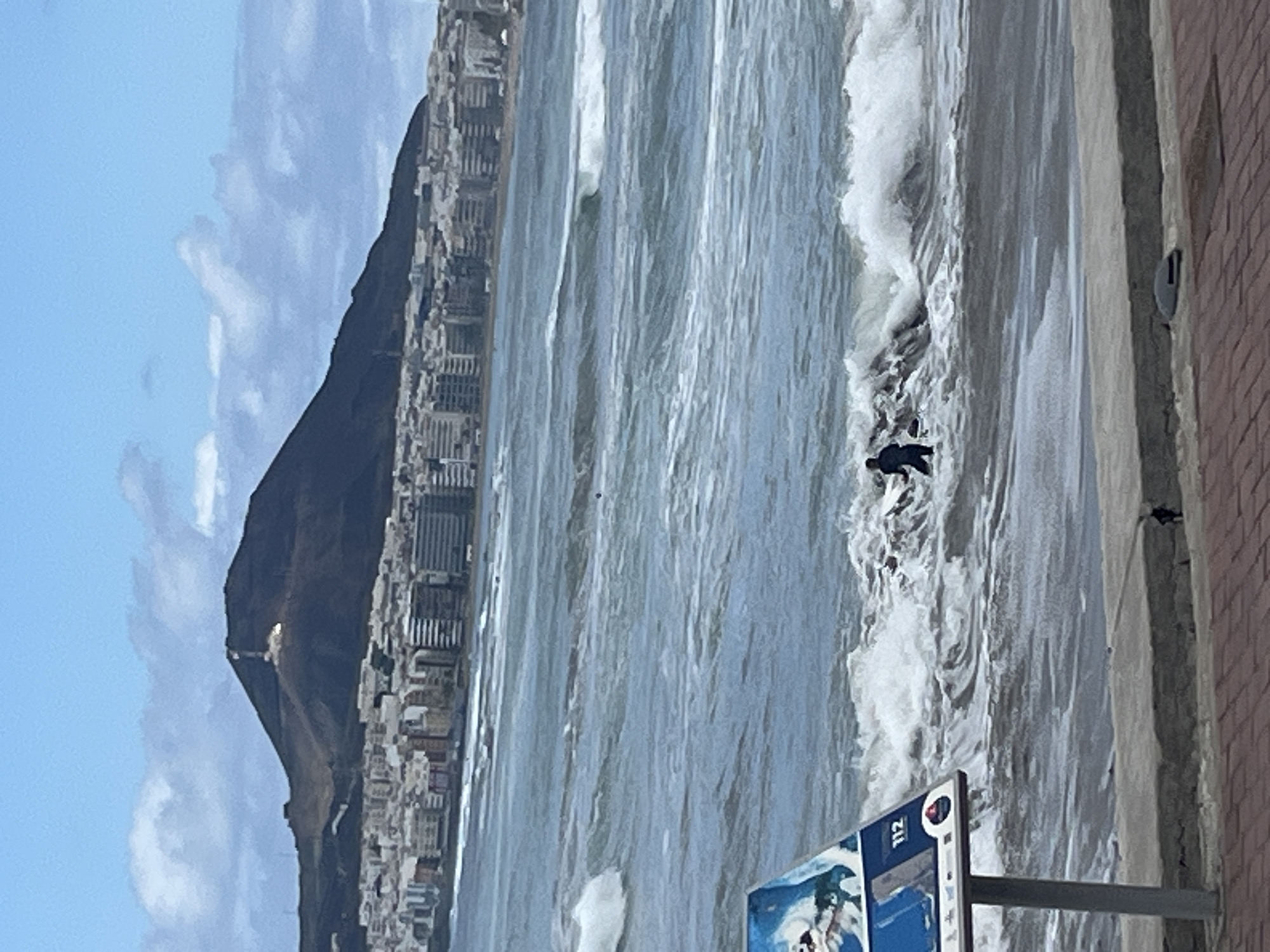 An outdoor, slightly tilted, eye-level shot captures a dynamic coastal scene in Las Palmas De Gran Canaria, Spain, on a partly cloudy day. The ocean dominates the left half of the frame, characterized by numerous blue-grey waves, many breaking into white foam as they approach the shore. A person, clad in a dark wetsuit, is visible in the foreground amidst the white foam of a breaking wave, appearing to be swimming or bodyboarding near the water's edge.
To the right, a concrete promenade or barrier separates the viewer from the water, with a brown brick wall running parallel to the coast. A blue and white sign, possibly an informational or emergency services sign, stands upright on a pole in the bottom right corner, displaying the number "112" prominently.
In the background, a large, dark mountain dominates the skyline, its base covered with numerous white and grey buildings that stretch along the coastline. A small, bright, rectangular structure or reflection is discernible on the upper slope of the mountain. The sky above is a light blue with scattered white clouds. The overall impression is one of a bustling city coastline with active ocean conditions.