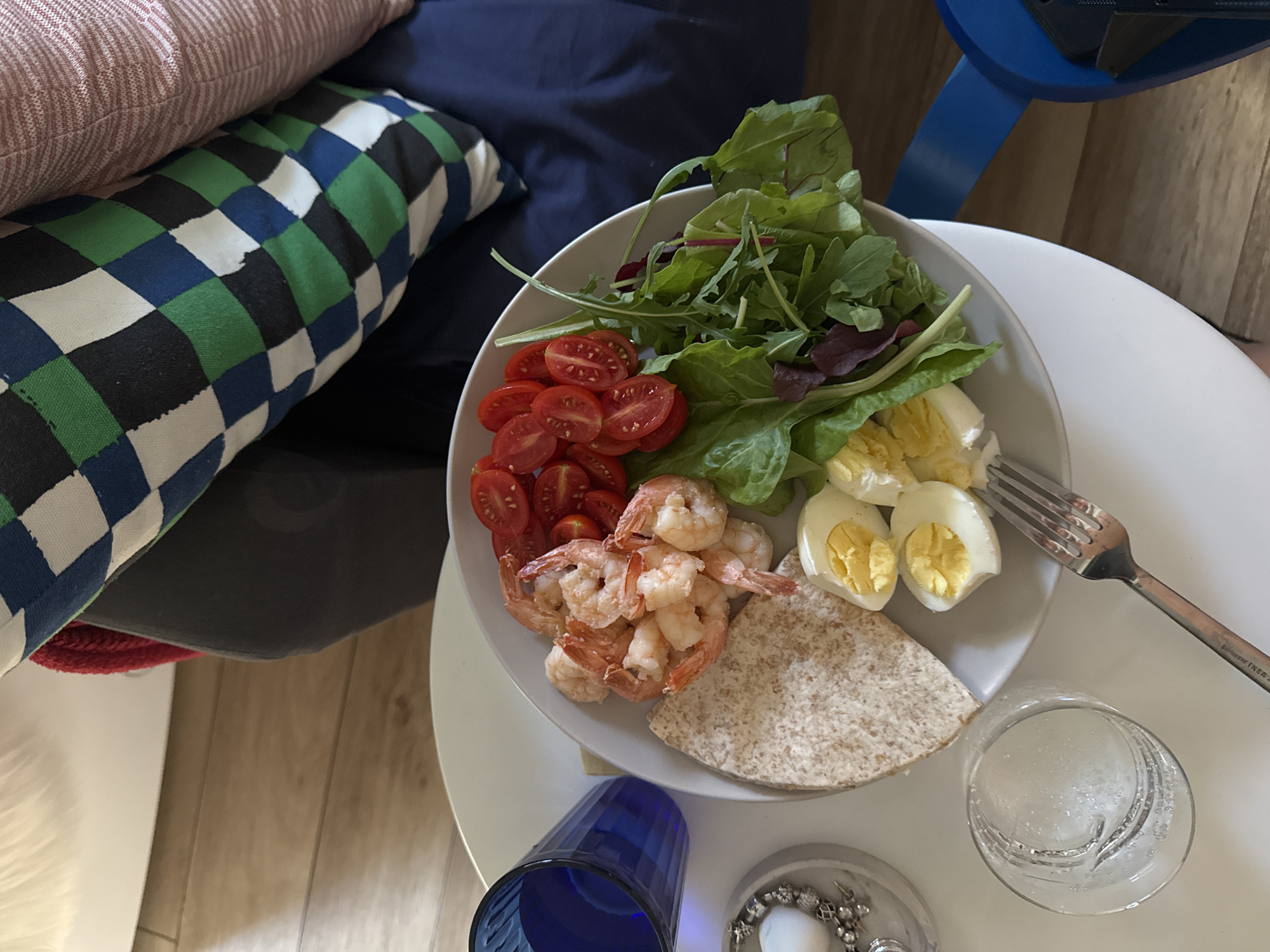 A plate of food rests on a white circular table. The plate contains cooked shrimp, halved cherry tomatoes, a portion of whole wheat flatbread, a salad of mixed greens, and sliced hard-boiled eggs. To the left of the plate is a blue glass tumbler filled with a clear liquid, and a small white bowl containing a candle and jewelry. A silver fork rests on the table in front of the plate. In the background, out of focus, are a sofa with a checkered pillow and a wooden floor. This image was taken in Tel-Aviv, Israel.