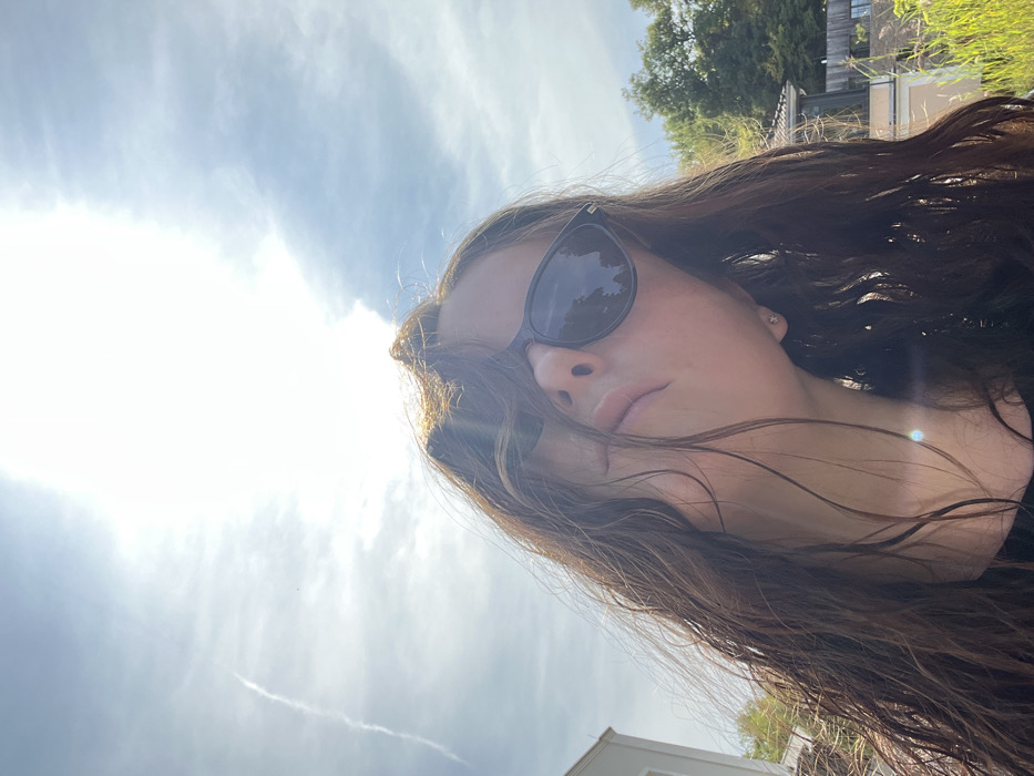 A low-angle, upward-facing photograph captures a subject, likely female, with long, wavy, dark brown hair that appears damp, obscuring parts of her face. She wears dark-framed sunglasses with reflective lenses, partially revealing her nose and lips. A small, reflective stud earring is visible in her right earlobe. The upper portion of the image is dominated by a bright, partly cloudy sky with a prominent sun glare near the top center, and a faint contrail is visible in the upper left. In the background, on the left, a portion of a light-colored modern building is visible. On the right, beyond green foliage, a building with a reddish-brown tiled roof and light-colored walls is partially seen, alongside tall green grass or reeds. The scene is situated in Bad Sobernheim, Germany.