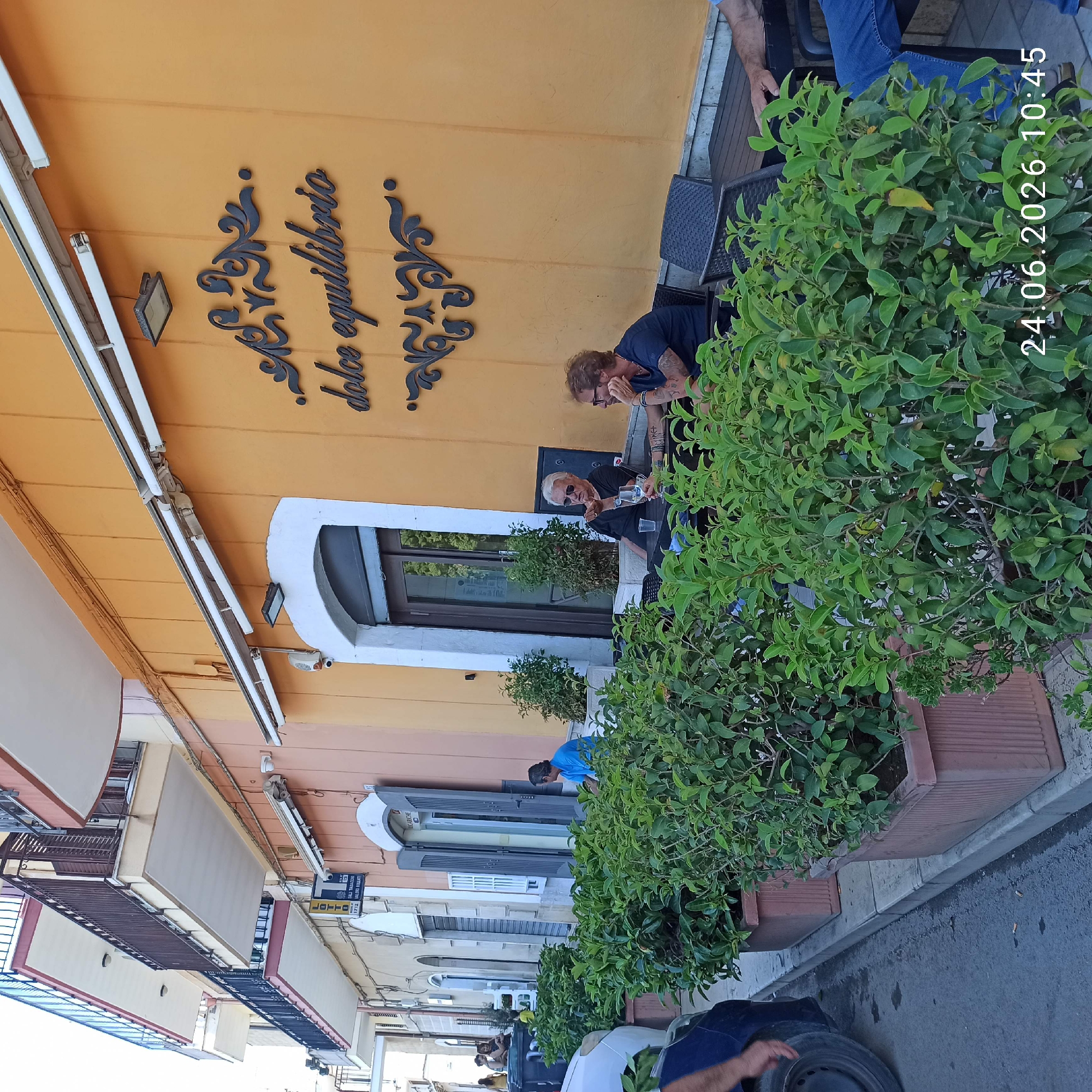 In Bitonto, Italy, on June 24, 2026, at 10:45 AM, an outdoor seating area of a cafe is depicted. The cafe's facade is painted orange with decorative dark brown script signage that reads "dolce equilibrio." Two men are seated at a dark table. The man on the left, with white hair and sunglasses, wears a black t-shirt and holds a glass. The man on the right, with reddish-brown curly hair and glasses, wears a navy blue t-shirt and has tattoos on his arm. Both men appear to be engaged in conversation. The foreground features several large planters filled with lush green foliage, suggesting a street-side cafe. A portion of a car's tire and fender is visible on the left edge of the frame. In the background, there are other people sitting and walking on a sidewalk, with buildings featuring balconies and architectural details lining the street. The overall scene captures a moment of relaxation at a cafe in an Italian town.