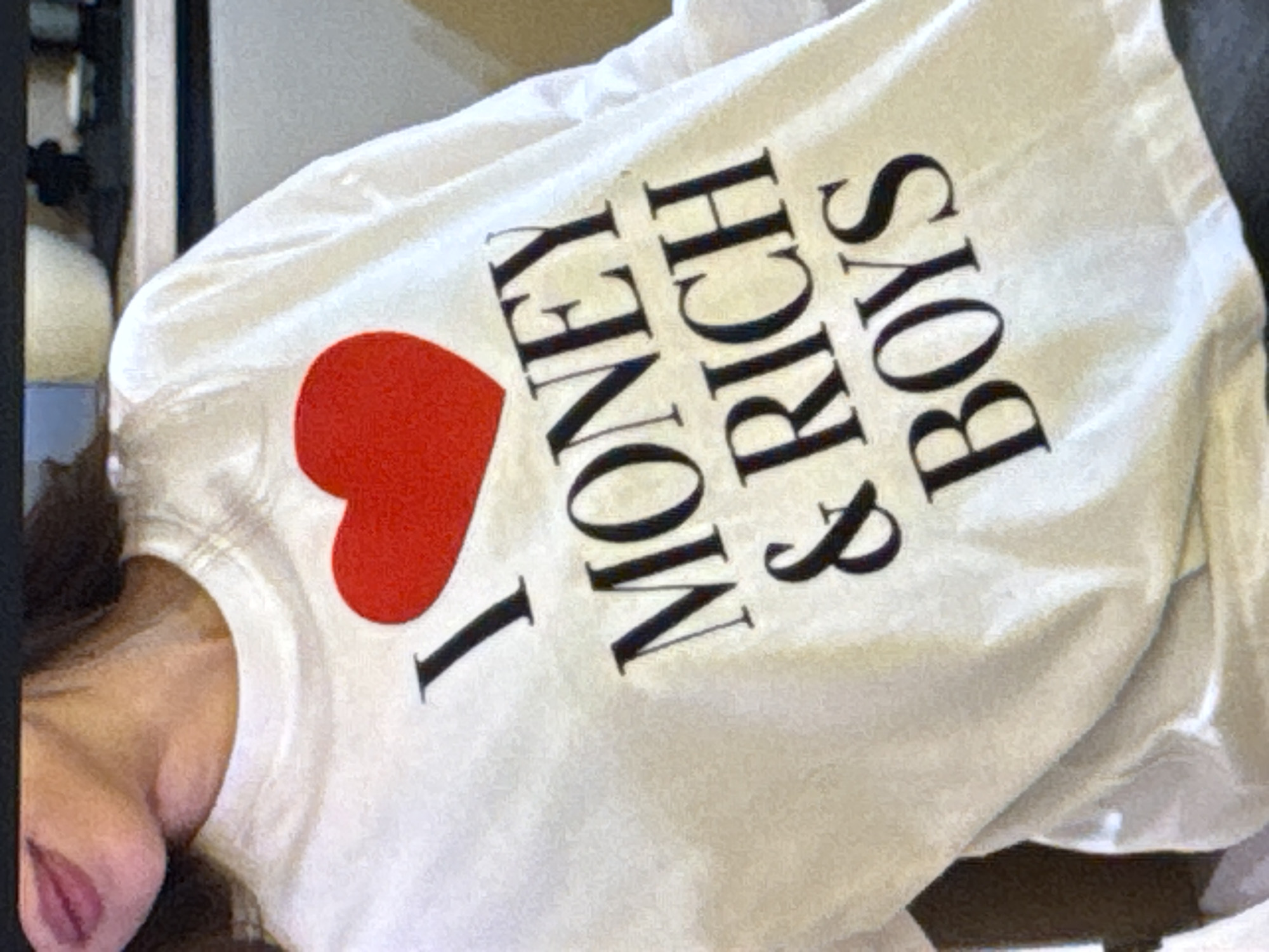 The media file shows a white t-shirt with a graphic design. The design consists of a red heart symbol followed by the text "I MONEY & RICH BOYS". The t-shirt is worn by a person, with their shoulder and part of their neck visible in the lower left portion of the frame. The fabric of the t-shirt is slightly wrinkled, suggesting it might be laid flat or casually worn. The lighting in the image is bright, indicating it was likely taken indoors with good illumination or outdoors on a sunny day. There are no strong environmental cues to suggest a specific time of day or weather conditions beyond general brightness. The background is mostly out of focus, with some dark vertical elements and lighter, indistinct shapes, offering little information about the setting. The focus is clearly on the t-shirt and its graphic. The overall impression is casual, with the prominent text on the t-shirt being the main point of interest. The location context of Kuta Selatan, Indonesia, does not provide any specific visual information that can be deduced from the image itself.