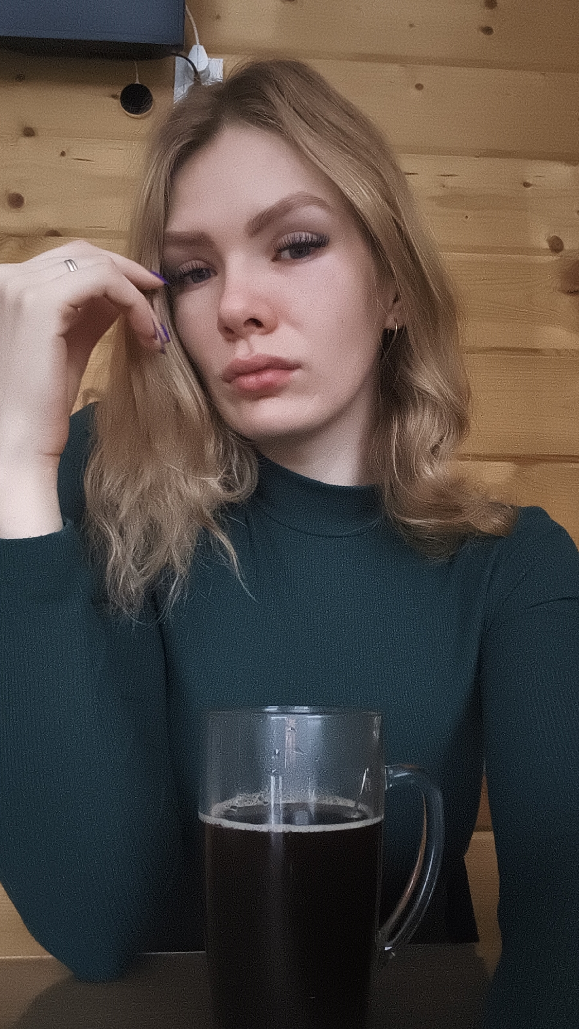A young woman with blonde, wavy hair is sitting indoors, posing for a selfie. She's wearing a dark green, long-sleeved top. Her left hand is raised, fingers touching her forehead, and she is wearing a ring on one of her fingers with violet painted fingernails. In the foreground, there is a large glass mug filled with a dark liquid, possibly beer or coffee. The background consists of wooden paneling, possibly a wall, with what appears to be a black electronic device mounted at the top left corner of the frame. There is no indication from the image of the specific location in Русский Вожой, Russia.