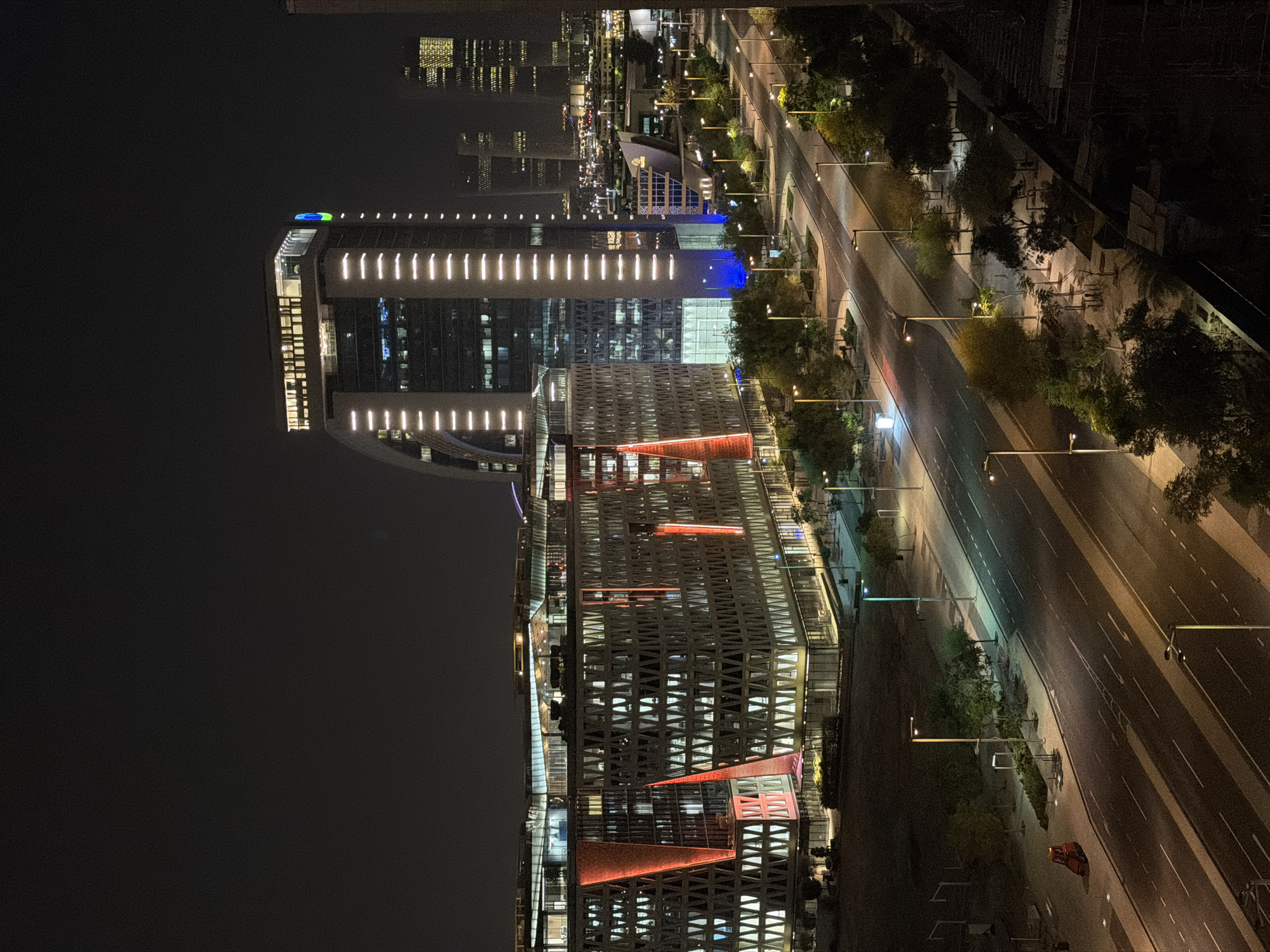 The image shows a nighttime view of a modern cityscape. In the foreground, a street with multiple lanes is illuminated by streetlights. There are trees and sidewalks lining the street. On the left side of the street, there is a large, modern building complex with geometric patterns and red-colored accents. Further in the background, another tall, modern building with vertical white lighting stands out, and beyond that, more skyscrapers with illuminated windows are visible. The sky is dark, indicating it is nighttime. A small, red maintenance vehicle is parked on the side of the road in the lower left quadrant of the image. The overall scene depicts a well-lit urban environment at night, characterized by contemporary architecture and infrastructure.