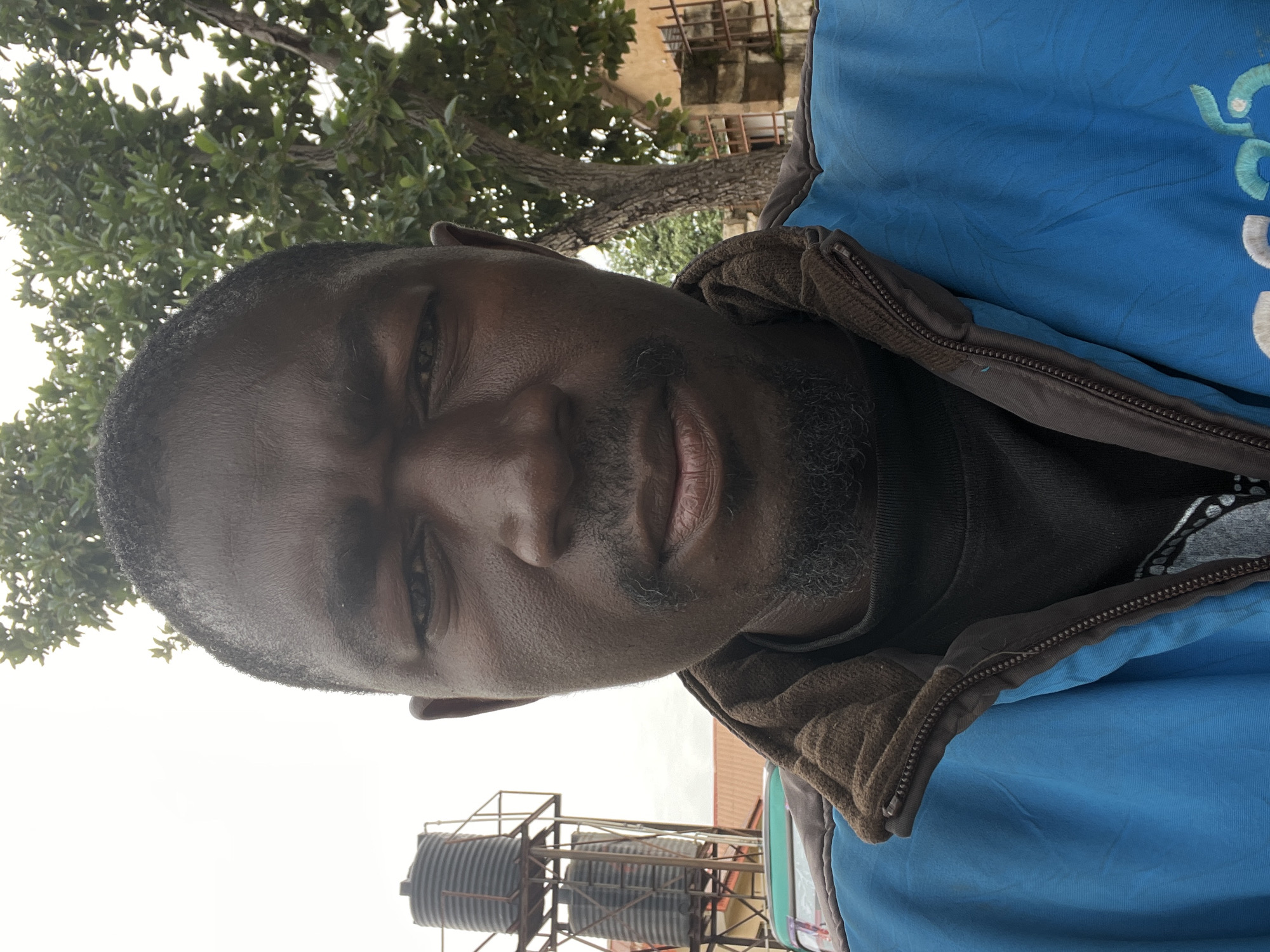 A man of African descent, with dark skin, a short beard, and dark hair, looks directly at the camera. He is wearing a blue jacket with a brown collar and a dark t-shirt underneath. Behind him, a tall, multi-tiered water tank stands against a pale sky. A large tree with green leaves is to the right of the tank. In the background, parts of buildings and a fence are visible. The scene appears to be in Jos, Nigeria.