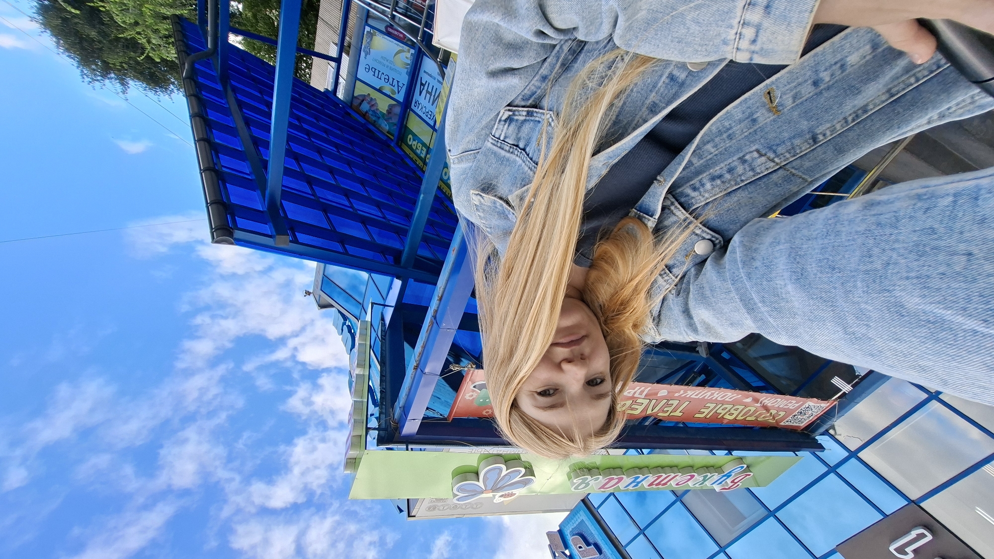 This is a selfie of a young woman with blonde hair, wearing a denim jacket and jeans. She is outdoors, with a blue sky and white clouds in the background, suggesting it is daytime and fair weather. The setting appears to be a commercial or public area, with a building featuring blue paneling and a transparent roof structure.
There are several signs visible, though some are partially obscured or difficult to read due to the angle and resolution. One sign, in red and white text, appears to contain Russian words like "РЕМОНТ," "ДОКУМЕНТЫ," and "ТЕПЛЫЕ." Another sign, in green and white, has Cyrillic text and a stylized flower graphic, possibly indicating a business or service. The building's architecture suggests a modern design. The overall mood conveyed by the selfie is casual and relaxed. The image is oriented sideways.