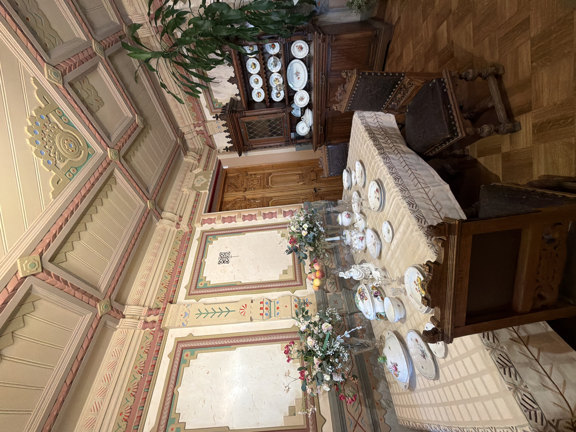 The image depicts a dining room interior, possibly within a historic building in Nizhny Novgorod, Russia. The room features ornate detailing on the ceiling with a pink and cream color scheme. The walls are adorned with decorative panels and friezes. A large wooden dining table is set with a patterned tablecloth and a collection of porcelain dishes, including plates, bowls, and serving tureens. Two floral arrangements are placed on the table. To the right, a dark wooden hutch or cabinet displays a collection of plates with fruit motifs on its shelves. Two antique wooden chairs with dark upholstered seats are positioned at the table. In the background, a wooden door with intricate carvings is visible. The floor is made of dark parquet flooring. A large green plant is situated in the corner of the room. No people are present in the scene.