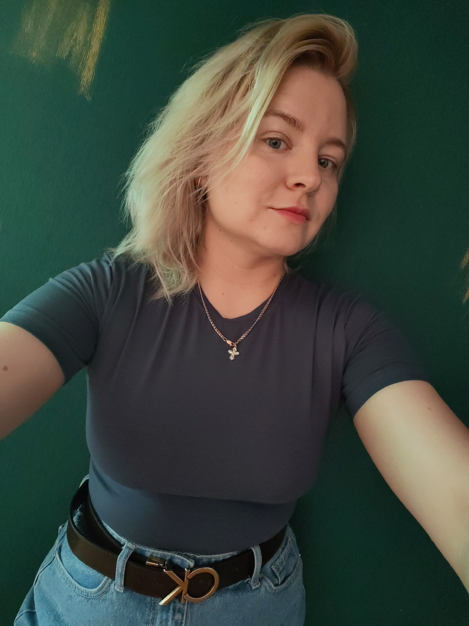 A young woman with blonde hair and blue eyes is looking at the camera, leaning against a dark green wall. She is wearing a dark grey t-shirt, blue jeans, and a black belt with a gold buckle that appears to be a Calvin Klein logo. A silver necklace with a cross pendant is visible around her neck. The lighting suggests an indoor setting, possibly a room with a painted wall. There are some lighter streaks on the wall in the upper left portion of the image, which may indicate artistic brushstrokes or a different texture. The overall composition is a close-up shot, focusing on the woman's face and upper body. The specific location in Izhevsk, Russia, cannot be determined from the provided image.