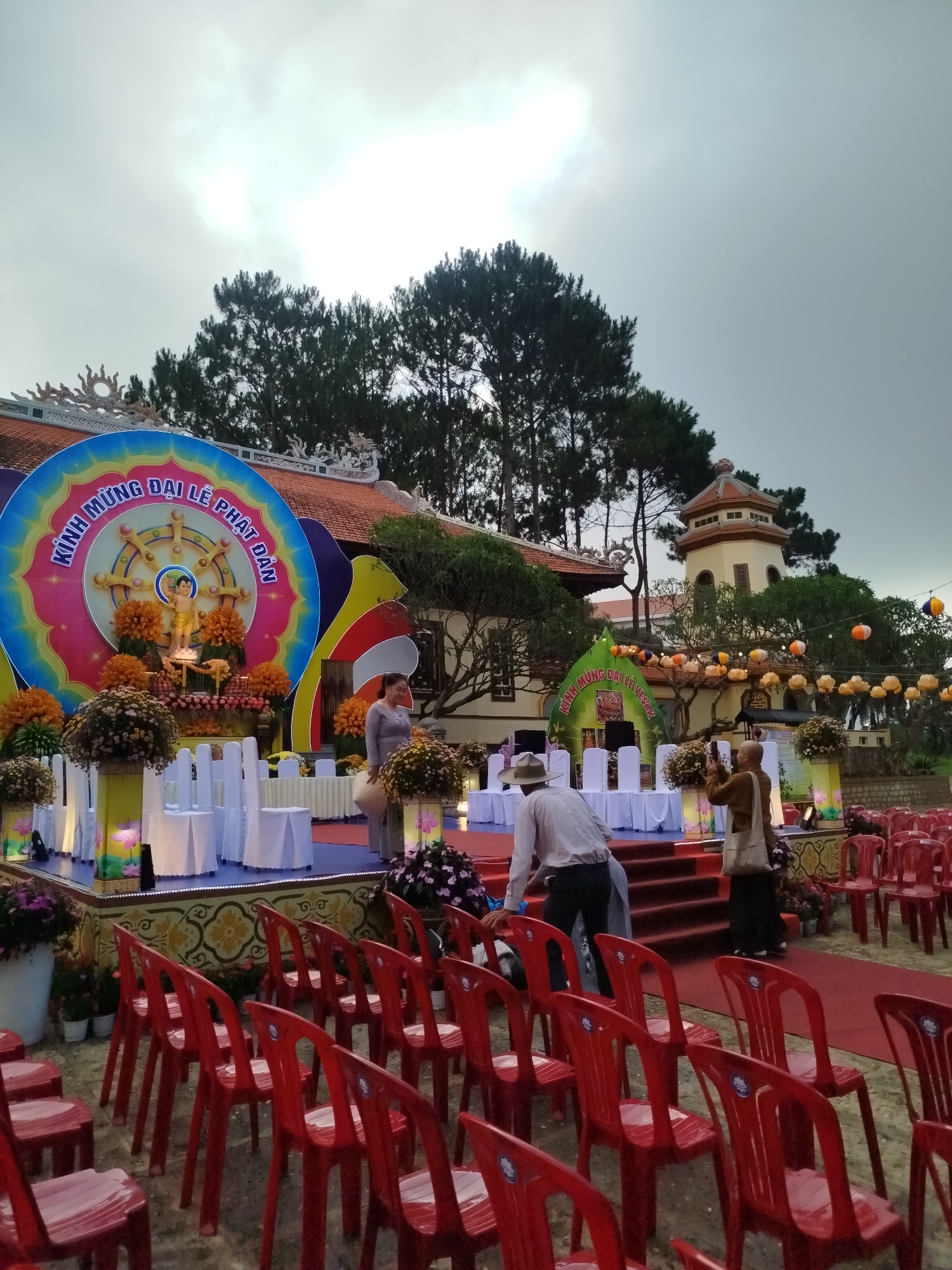 The scene depicts preparations for a religious event in what appears to be a temple complex in Da Lat, Vietnam. In the foreground, rows of empty red plastic chairs are arranged, suggesting seating for attendees. A red carpet leads towards a stage decorated for the event.
The stage is adorned with colorful backdrops featuring religious iconography and the text "KINH MỪNG ĐẠI LỄ PHẬT ĐẢN". Statues and floral arrangements are displayed on the stage, while white tablecloths cover the tables and chairs.
Several individuals are present: a woman in a grey dress stands near the stage, a man in a white shirt and hat walks towards the stage, and another person, possibly a woman, wearing a brown outfit, stands on the stairs, taking a photo with her phone.
Behind the stage, the architecture of the temple complex is visible, including a multi-story tower and a building with a red tile roof. Lanterns are strung between the buildings, adding to the festive atmosphere. Large trees stand in the background.
The overcast sky suggests that the weather is cool, a characteristic often associated with Da Lat.