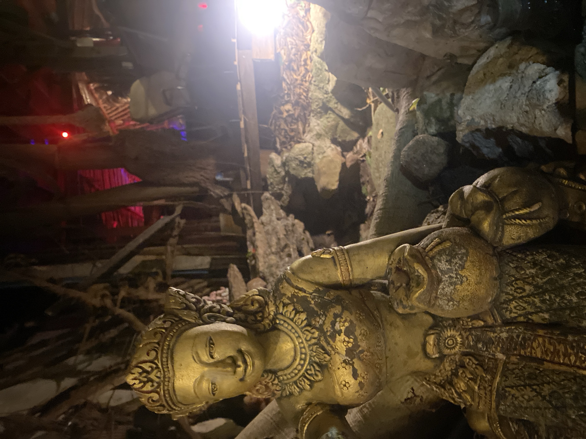 A gold statue is located in Baan Don Sai, Thailand. The statue is situated in a cave-like environment with rocks and root-like structures surrounding it. The area above appears to be a constructed ceiling or overhead structure with red and white lights.