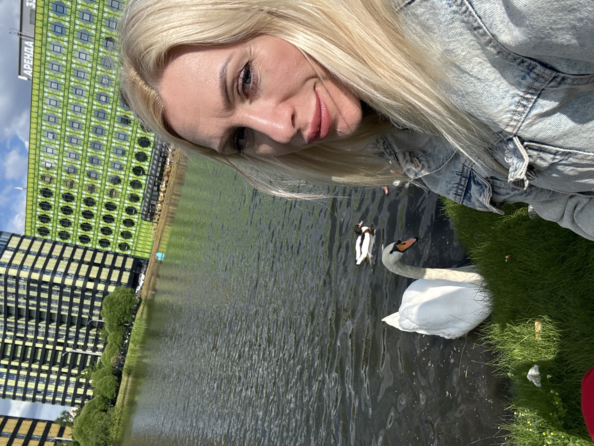 A woman with blonde hair, wearing a denim jacket, takes a selfie by a body of water. In the water, a white swan and a duck are visible. Behind the water, there are modern buildings with yellow and green facades. One building has "АРЕНДА" (rent) written on it. This scene is located near the park in the residential complex "Golden City" in Saint Petersburg, Russia, a coastal area along the Gulf of Finland, a district known for its modern architecture.