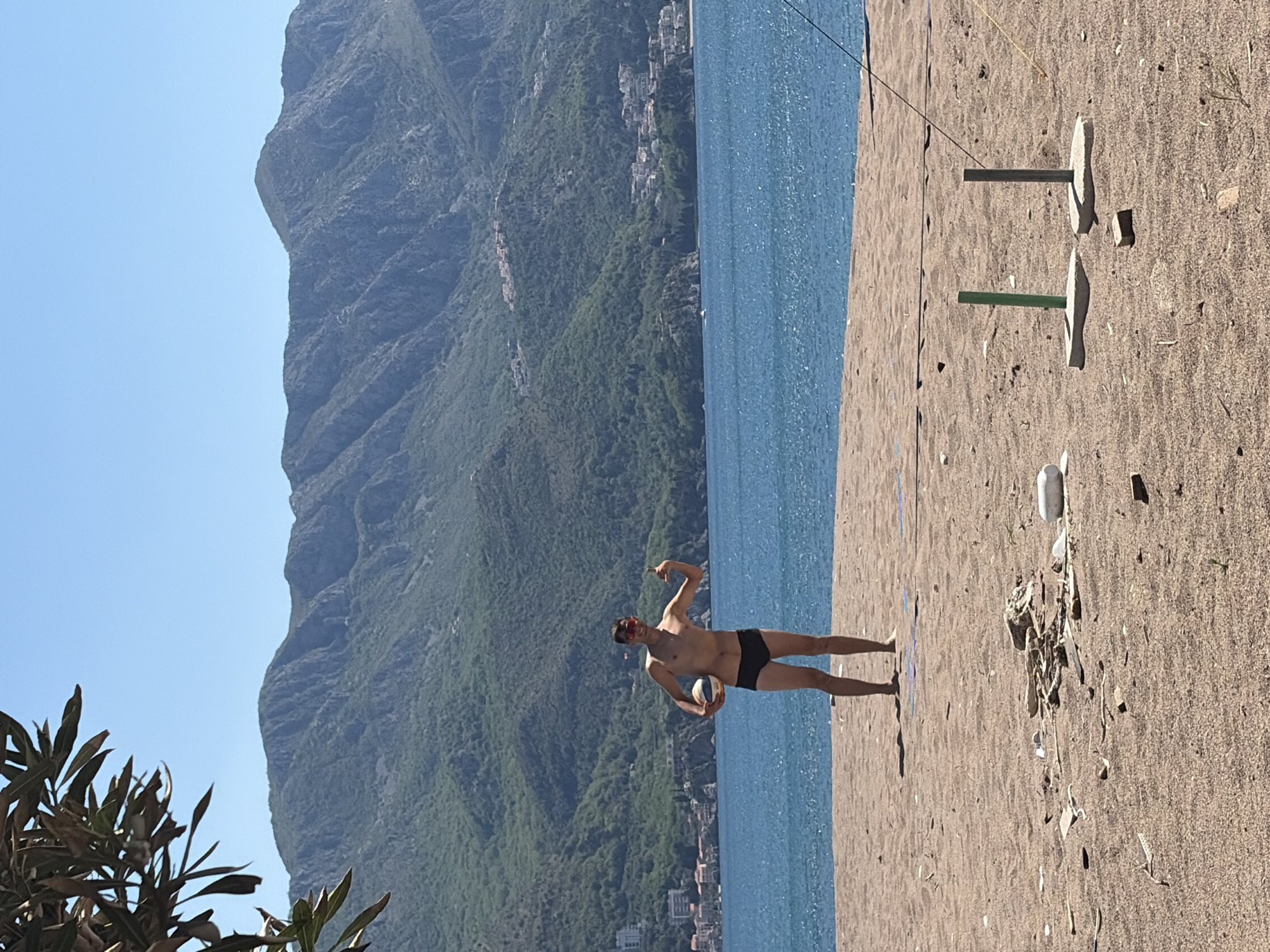 The image shows a man holding a volleyball on a beach. The man is wearing sunglasses and black shorts. A beach volleyball net is visible behind him. The sea is in the background, with mountains and buildings further in the distance. The location appears to be Bečići, Montenegro.
