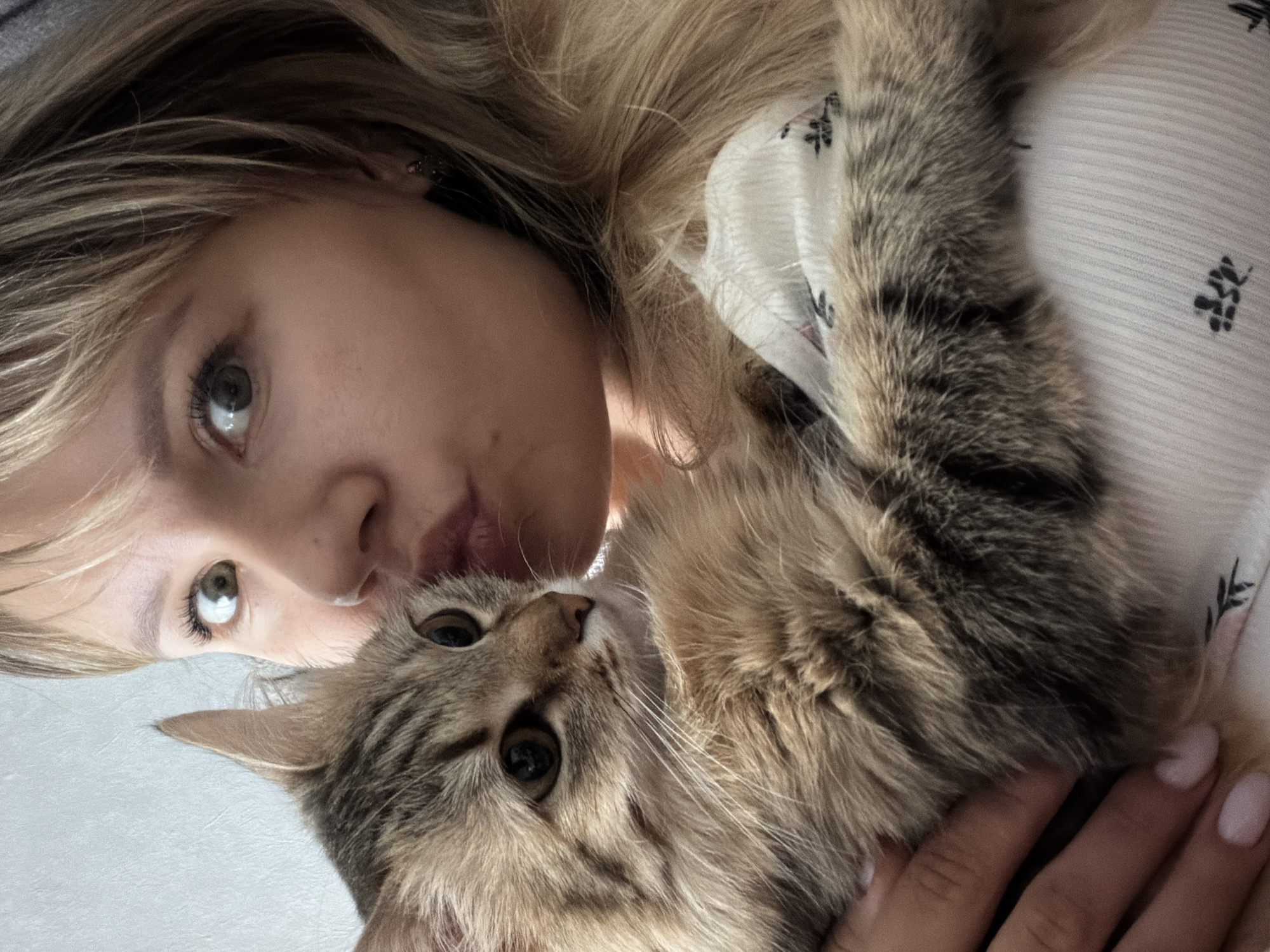 A young woman with blonde hair is holding a tabby cat. She is looking at the camera with a slight pucker of her lips. The cat is looking upward to the right. The woman is wearing a white shirt with a small black floral print. The background is a light grey or white surface. The location is unspecified and there is no indication that the image was taken in Chaikovsky, Russia.