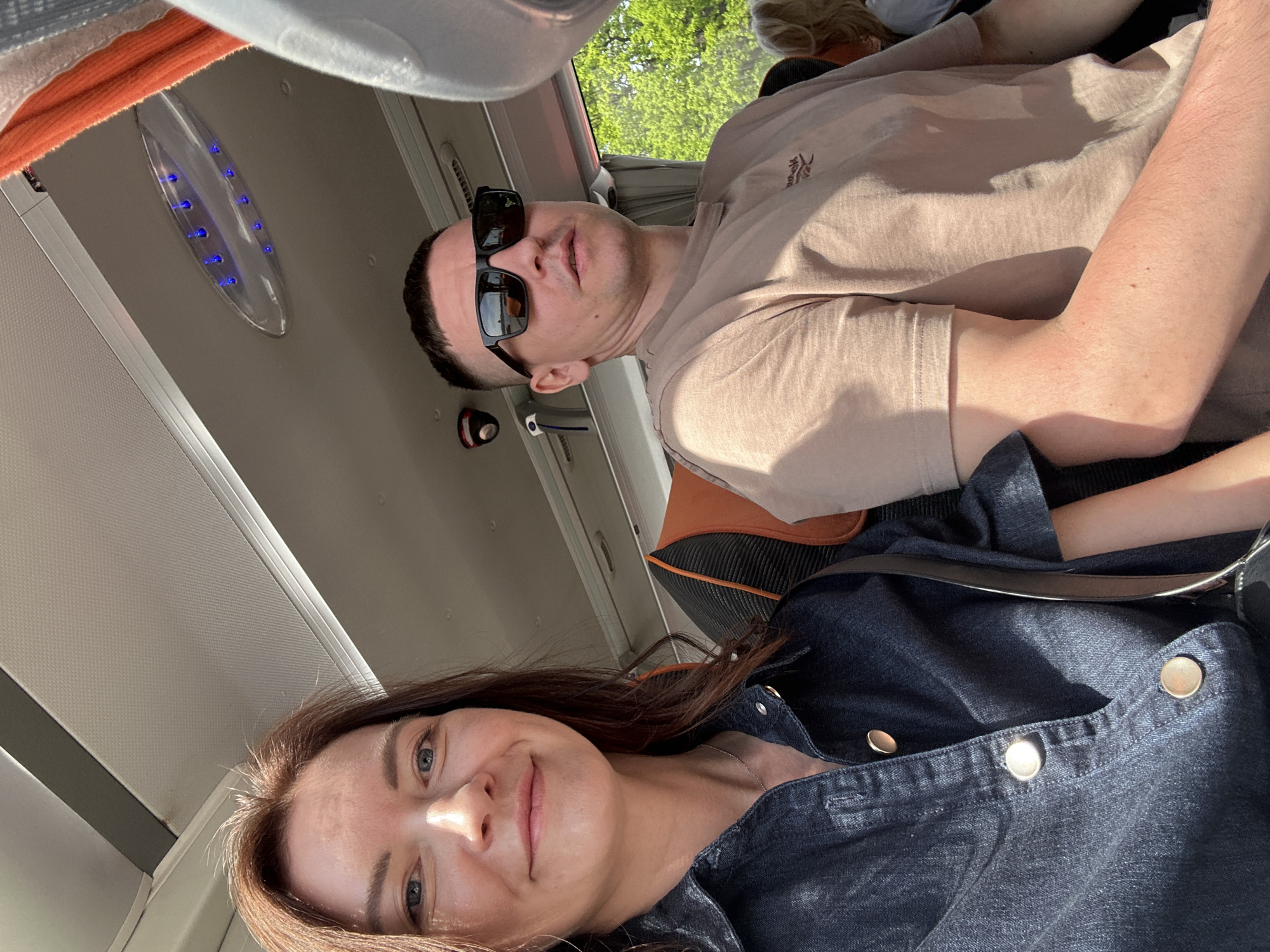 Two people are seated, likely on a bus or plane based on the overhead light panel and window placement. The man is wearing sunglasses and a light tan t-shirt. The woman is wearing a dark denim jacket with visible gold buttons. They are facing towards the camera, suggesting they may be taking a selfie. The location in the city of Unknown, Unknown is not determinable from the image.