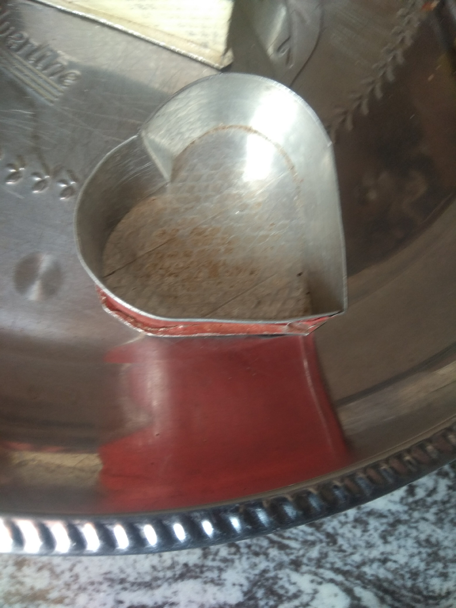 The image shows a close-up, overhead shot of a silver heart-shaped cookie cutter placed on a silver tray. The cookie cutter has a red rim and appears to be made of metal. The inside of the cookie cutter is filled with a light-colored granular substance, possibly flour or sugar. The tray has a decorative rim with a pattern of indentations. There is a hint of a red surface beneath the cookie cutter, reflecting on the tray. The background is out of focus, but suggests a textured surface. The image does not provide any information about a specific location in Jalingo, Nigeria, or any people or actions.