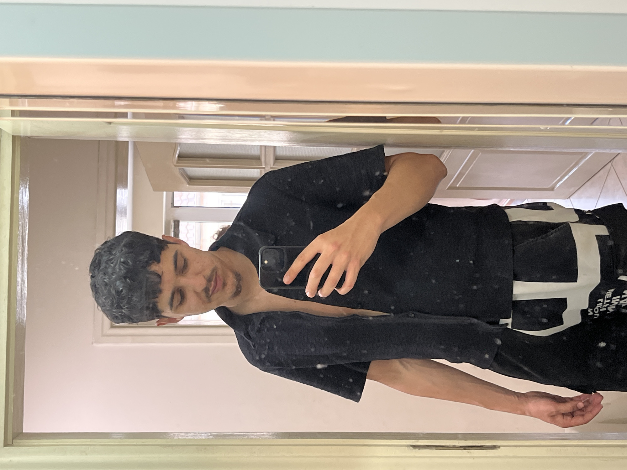 A young adult male, with dark curly hair and a fringe, dark mustache, and goatee, is depicted in a mirror selfie within an indoor setting in Varna, Bulgaria. He is wearing a black, short-sleeved, collared shirt, unbuttoned to reveal his chest, and black shorts or pants with large white graphic elements. He holds a dark-colored smartphone, likely an iPhone, in his right hand, positioned to capture his reflection. The mirror surface exhibits numerous small, light-colored specks. The background, visible in the mirror's reflection, consists of light-colored walls, a light-beige paneled door, and beyond it, a window revealing rooftops, suggesting an urban environment. A tiled floor is also reflected. The mirror is framed by light-colored elements, including a pale yellow/beige section on the left and top, and a wider pinkish-beige section on the right, adjacent to a pale blue wall or frame.
