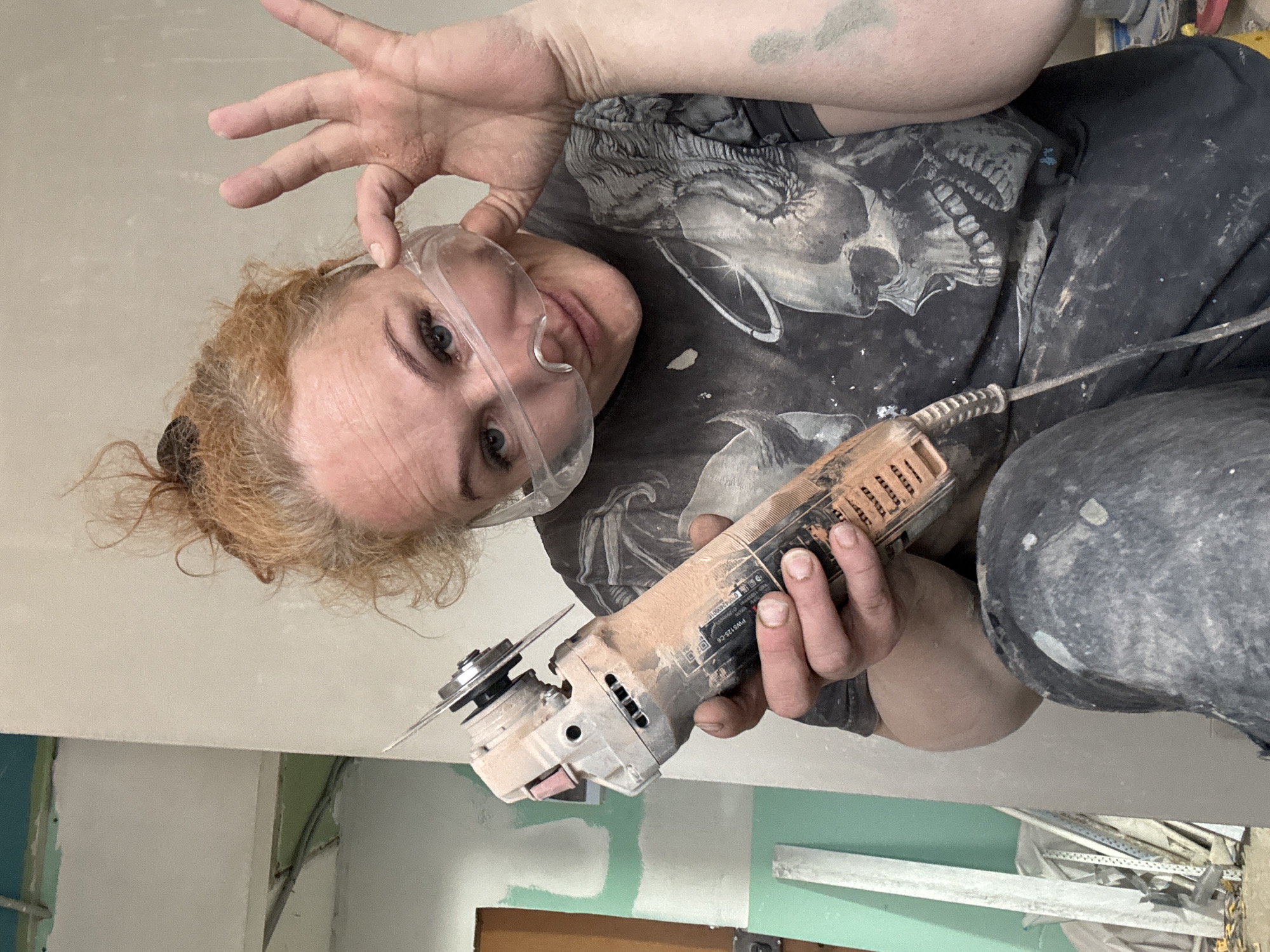 A woman is indoors, likely in the city of Chaikovsky, Russia, holding an angle grinder. She is wearing safety glasses, a dark t-shirt with a skull graphic, and dark trousers. Her red hair is pulled back. Her hands and clothing are covered in dust. The background shows unfinished interior construction, with green and white plaster.