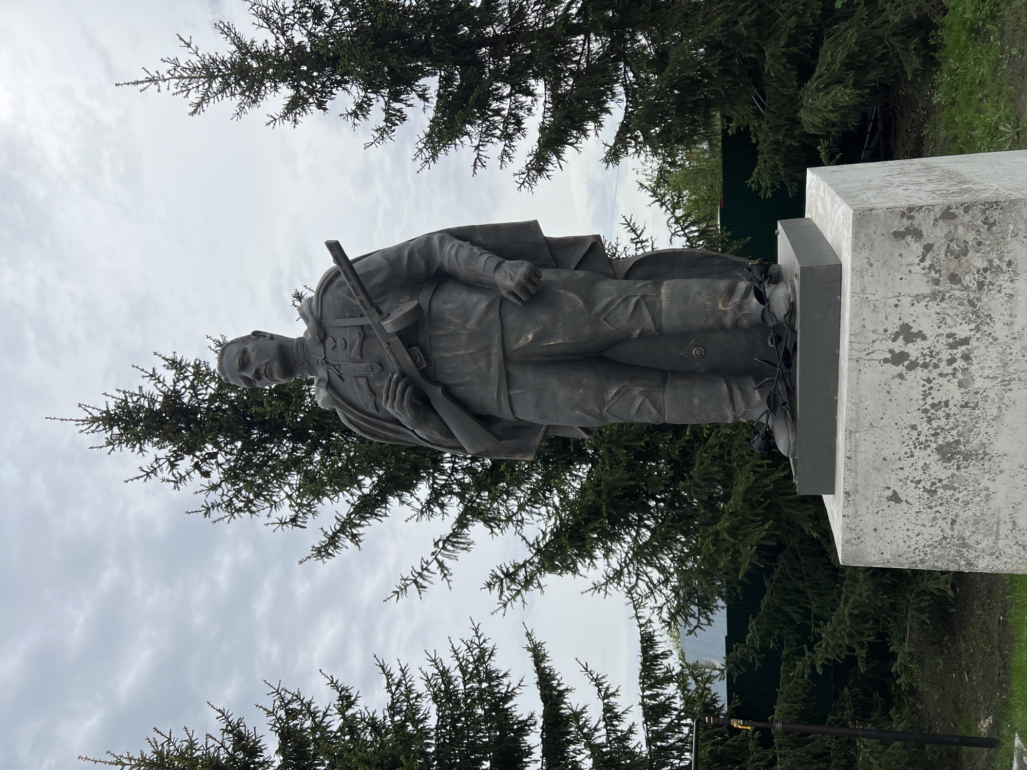The image depicts a bronze statue of a soldier standing on a concrete pedestal, likely a war memorial. The soldier is wearing a uniform and holding a weapon. The statue is surrounded by evergreen trees. The sky is overcast. The location is Введенская Слобода, Russia.