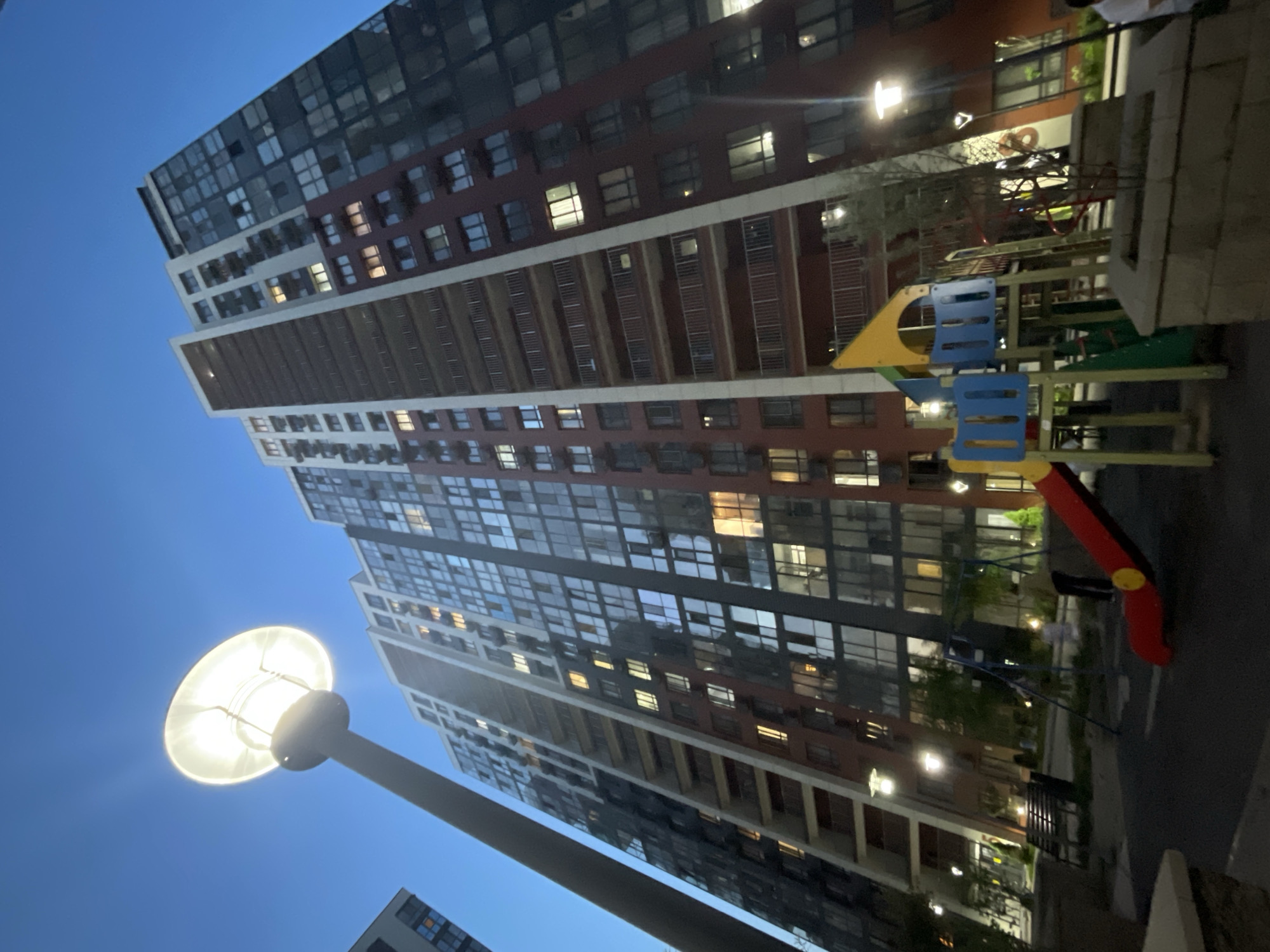 In Almaty, Kazakhstan, an exterior shot captures a modern residential high-rise building complex illuminated by streetlights at twilight. The sky is a deep blue, indicating dusk. A prominent streetlight with a large, circular lamp is positioned in the foreground, its light illuminating the surrounding area. To the right of the streetlight, a playground with a red slide, yellow and blue climbing structures, and swings is visible. The complex features tall buildings with numerous windows, some of which are lit from within, suggesting occupancy. Balconies with railings are visible on multiple floors. The overall scene depicts an urban residential setting after sunset, with recreational facilities and apartment buildings.