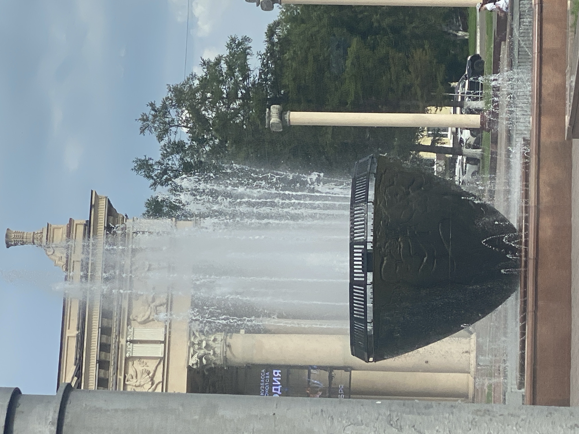 The image shows a fountain in front of a building with columns. There are trees in the background and a few cars parked on a street. A person is sitting on a bench in the distance. The fountain is large and elaborate, with water spraying high into the air. The building appears to be a historical or cultural site. The text on the building is in Russian and appears to be advertising a museum or exhibition. The image is taken on a bright, cloudy day. This scene is located in Novokuznetsk, Russia.