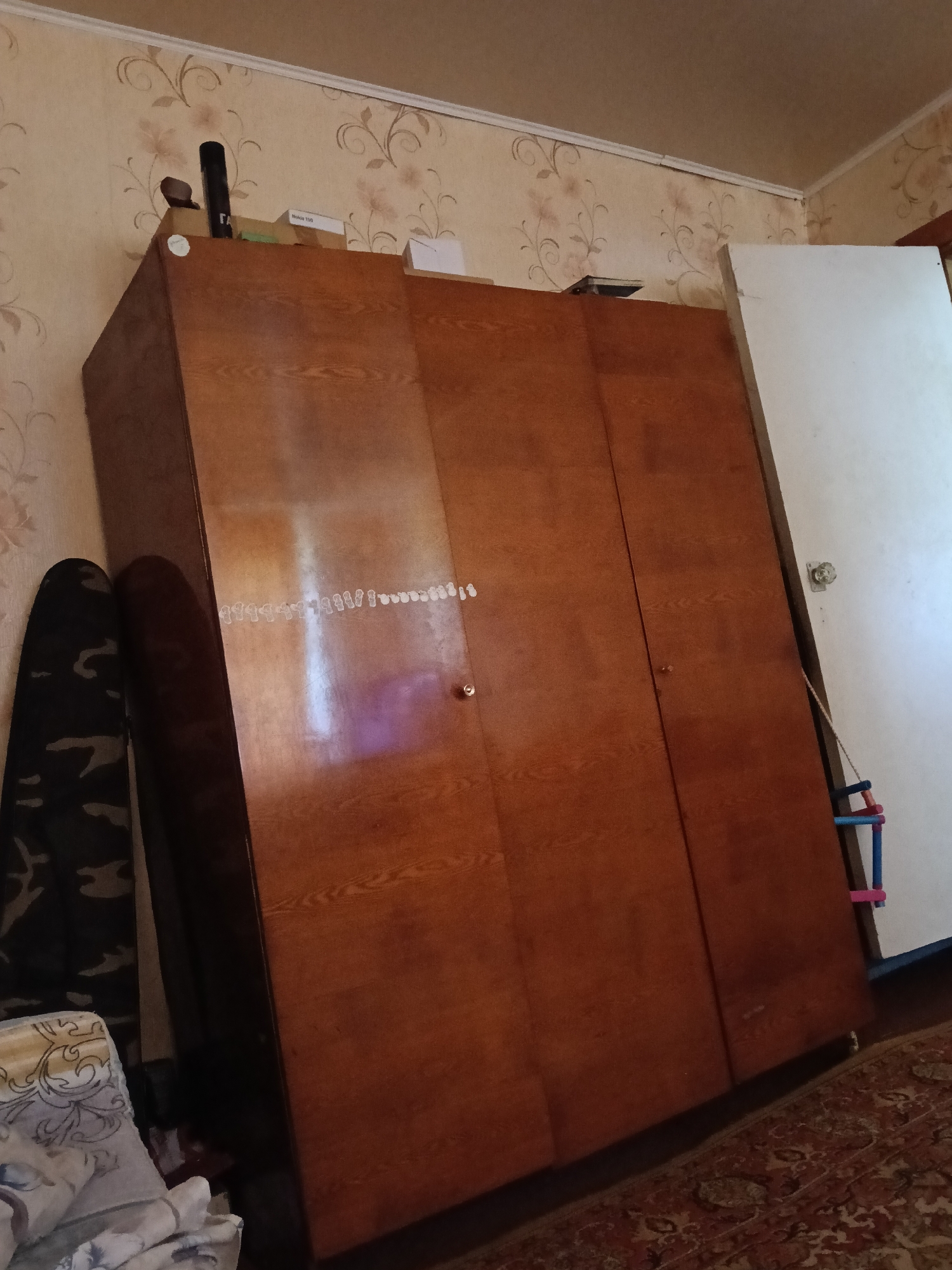 The image shows a cluttered room with a large wooden wardrobe dominating the center. The wardrobe has three doors and a light brown, wood-grain finish. A white door stands open to the right of the wardrobe. On top of the wardrobe, there is a black cylindrical object, a small brown object, and two cardboard boxes, one labeled "Nokia 100". A patterned rug covers the floor. On the left side of the image, a dark object with a camouflage pattern leans against the wardrobe. The walls are covered in floral wallpaper. The scene appears to be an indoor residential setting in Rameshki, Russia, suggesting a typical domestic environment.