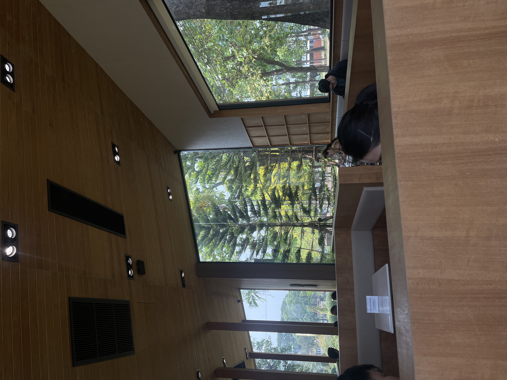 The image shows the interior of a building in Chiang Mai City Municipality, Thailand. The room features wooden walls, ceilings, and furniture, with built-in seating along a wall with large windows offering views of trees and greenery outside. Several people are present, with some sitting at the tables and appearing to be reading or working. The room has a modern design with embedded lighting fixtures and ventilation grilles. The overall atmosphere suggests a relaxed and contemplative environment.