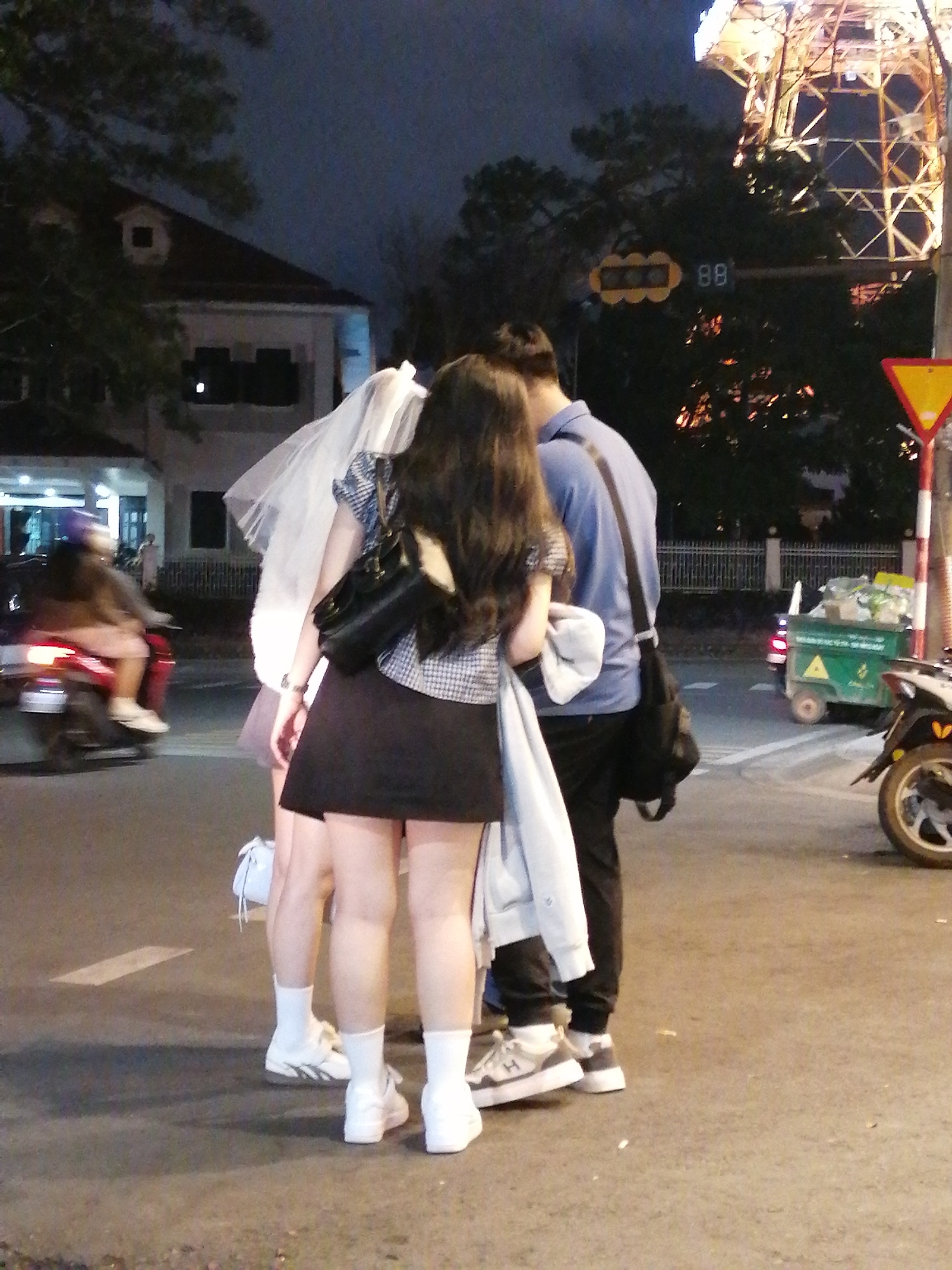 The image depicts a street scene at night in Da Lat, Vietnam. In the foreground, three individuals are partially visible, with two women and one man. One woman is wearing a black skirt, a checkered top, and a black handbag, with a white veil draped over her. The other woman appears to be wearing a light-colored skirt and white socks with sneakers. The man is dressed in a blue shirt, black pants, and a black bag slung across his body. They appear to be standing together on a street.
In the background, a red motorcycle with a rider is moving past on the left side of the image. Further in the background, a building with a dark roof and windows is visible, along with trees. A traffic light structure with illuminated signals and digital displays showing "88" is present. To the right, a yellow triangular warning sign on a pole is illuminated. A green refuse cart with wheels is also visible. The street surface is asphalt, with painted white lines. The overall lighting suggests it is nighttime, with artificial lights illuminating the scene.