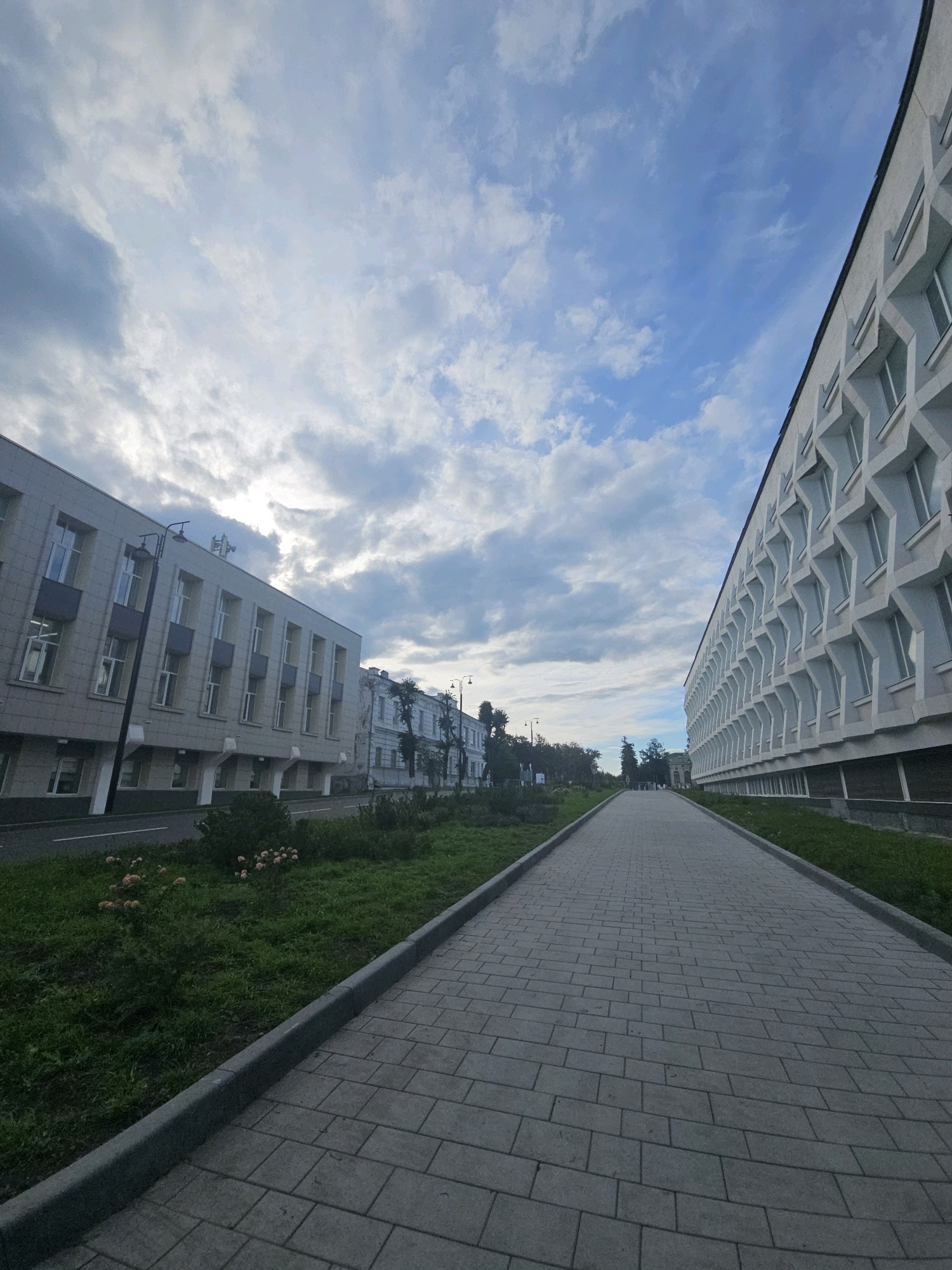 The image shows a wide, paved pedestrian walkway stretching into the distance. To the left of the walkway are grassy areas with some bushes and a street bordering them, adjacent to a multi-story building with numerous windows and a light-colored facade. Further down the street, another building with a white, textured facade and repeating geometric window designs is visible. To the right of the walkway, a similar building with the same distinctive window pattern continues along the side. The sky is partly cloudy with patches of blue. The scene appears to be an urban or institutional setting, possibly a university campus or a government complex, located in Ulyanovsk, Russia. There are no people visible in the frame. The lighting suggests it is daytime, but the clouds indicate overcast conditions or early morning/late afternoon light.