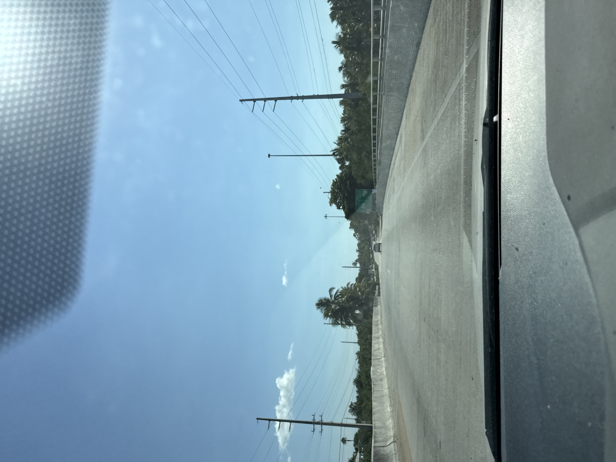 The image captures a view from inside a vehicle traveling on a multi-lane road. The road appears to be an elevated highway or bridge, with concrete barriers lining the sides. A row of palm trees and dense green foliage are visible beyond the barriers. Utility poles with numerous wires are interspersed along the landscape. The sky is clear and blue, with a few scattered clouds. In the distance, other vehicles are seen on the road. The overall environment suggests a tropical or subtropical setting. The perspective indicates the vehicle is moving forward, suggesting the scene is a snapshot of a journey.