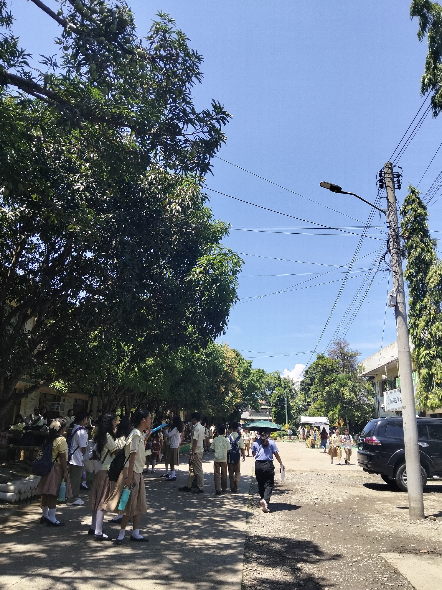 The image depicts an outdoor scene likely at a school in Davao City, Philippines, under a clear blue sky, suggesting it's midday. A group of students, predominantly in school uniforms consisting of light-colored tops and beige or brown skirts/trousers, are visible. Some are standing and conversing, while others appear to be walking. One student, wearing a blue shirt and dark pants, is walking away from the camera towards the right side of the frame.
Dominant elements in the scene include large trees with dense foliage, casting shadows on the ground, indicating a sunny day. Electrical poles with numerous wires crisscross the sky, a common sight in urban and semi-urban areas. A black SUV is parked on the right side of the unpaved road. In the background, buildings with signage are partially visible, including one with the text "DANIEL R. A."
The overall atmosphere appears to be that of a normal school day, with students gathering or moving around the campus. The bright sunlight and clear sky suggest good weather. The presence of many students in uniforms points to a school environment, possibly during a break, before or after classes.