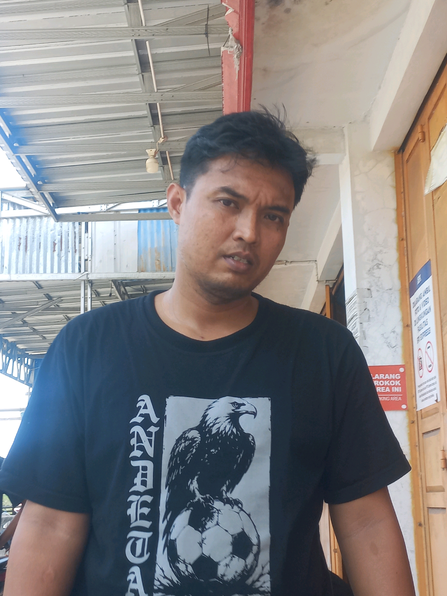 A medium shot captures a man with dark hair and brown eyes, looking directly at the camera with a neutral expression. He is wearing a black t-shirt with a graphic of an eagle perched on a soccer ball and the word "ANAKDAK" in vertical lettering. The man is positioned in front of a building with a corrugated metal roof and a wooden door. Signs are visible on the wall to the right, one reading "LARANG MEROKOK AREA INI" (No Smoking Area). The overall environment suggests an outdoor urban setting in Baturetno, Indonesia, likely a commercial or public space with simple architecture.