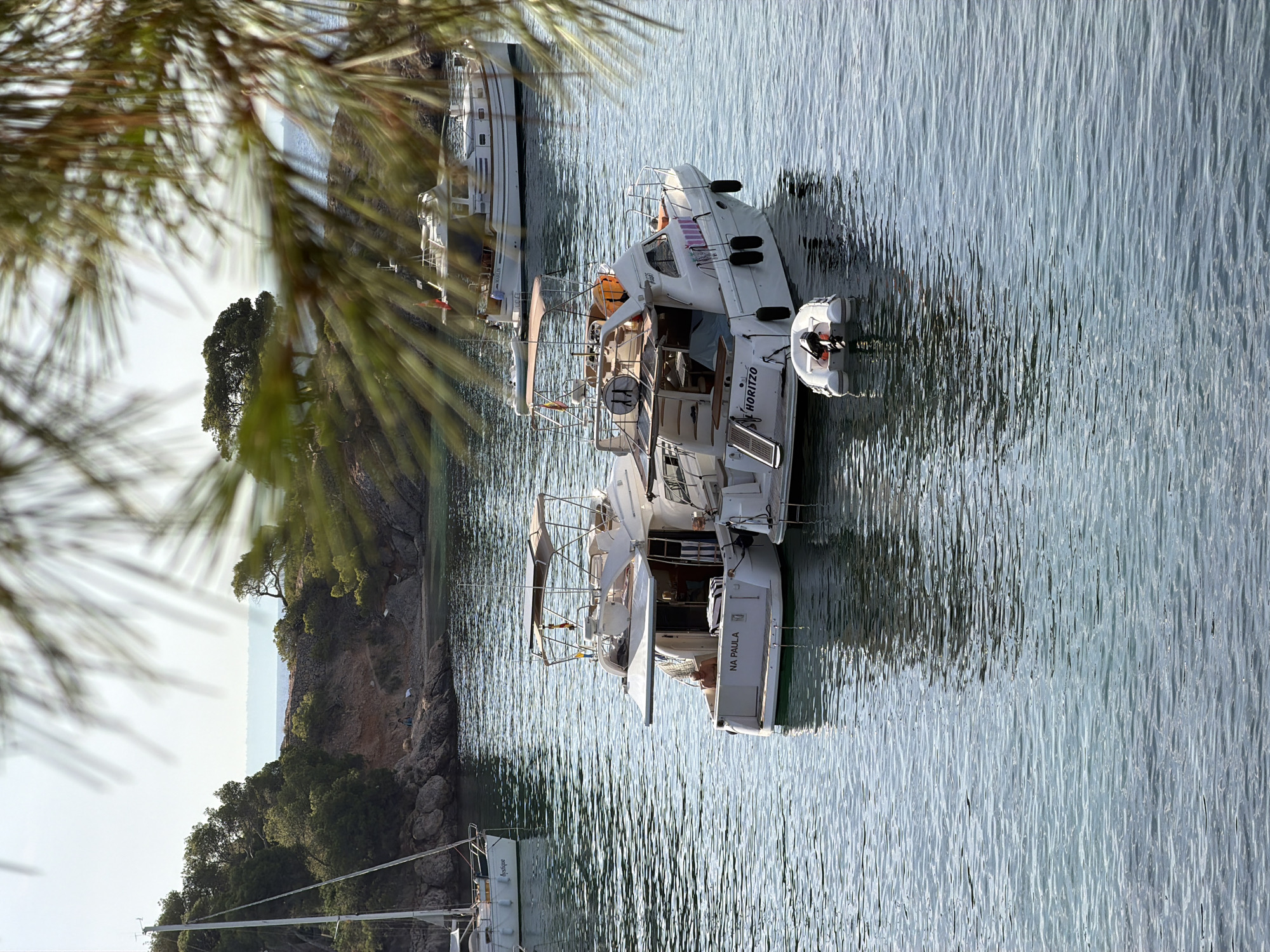 The image shows a body of clear, blue water with several boats moored. In the foreground, two white motor yachts are docked side-by-side. The yacht on the left has "NA PAULA" written on its stern. A smaller inflatable dinghy is attached to its stern. The yacht on the right has "HORITZO" written on its stern. Behind these yachts, further in the water, is another, larger white motor yacht with a dark hull trim. In the far left of the image, a white sailboat with a tall mast is visible, its hull partially obscured by foliage. The background features a lush, green, tree-covered hillside with rocky outcrops and a small beach. The sky is bright and hazy. The scene appears to be a tranquil harbor or bay, likely in a Mediterranean location given the vegetation and boat types. Based on the presence of Spanish-flagged vessels and typical coastal scenery, this location is consistent with the coastline of Calvià, Spain.