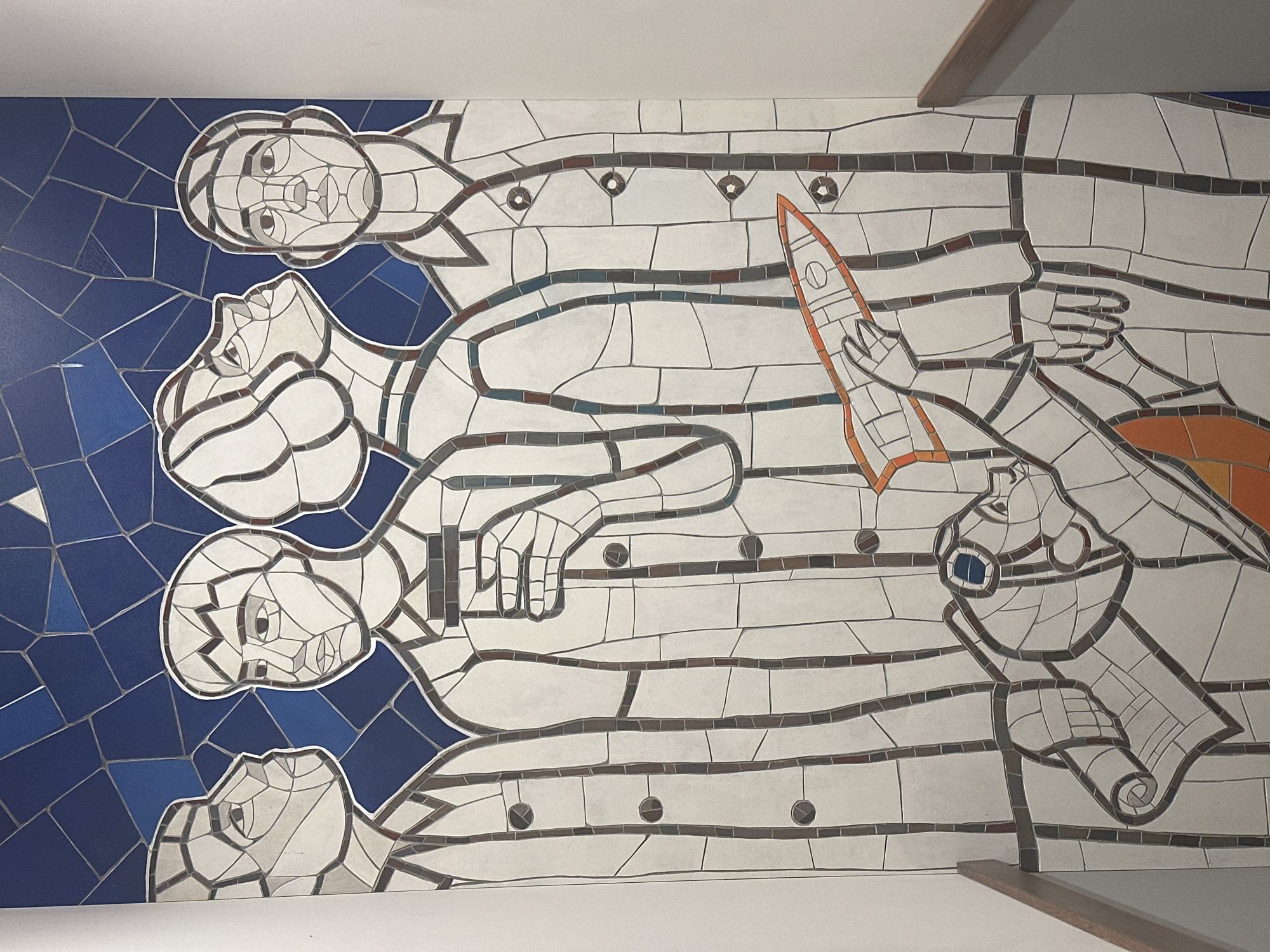 The image shows a mosaic mural located in Novosibirsk, Russia. The mural depicts four figures in a row, likely men, rendered in a stylized, mosaic-like pattern. The figures are primarily white with gray outlines and internal details, set against a blue background on the left that transitions to a white background on the right. The figures are oriented horizontally.
The top figure appears to be a man with a concerned expression. Below him is another figure, seemingly looking upwards. The third figure from the top is holding what appears to be an orange rocket in their hand. The bottom figure is holding a rolled-up document or blueprint, next to what appears to be a telescope or similar device.
The blue background on the left consists of geometric shapes arranged in a mosaic style. The overall impression is of a Soviet-era mural, possibly celebrating space exploration or scientific achievement. The style is distinctly graphic and stylized, with a limited color palette. The image is taken from a slightly low angle, showing part of the ceiling and a wooden beam.