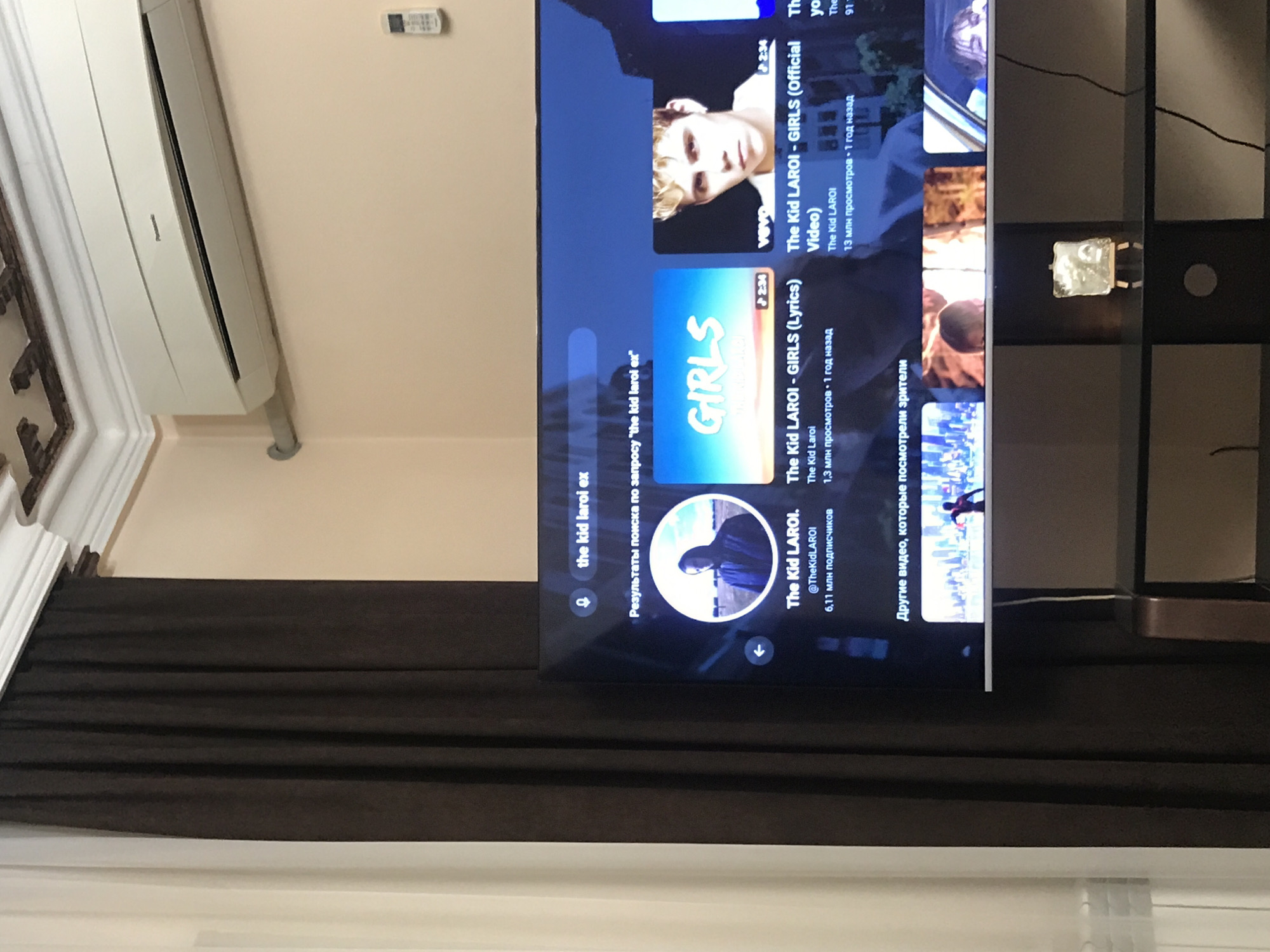 The image shows a modern interior with a focus on a television screen displaying search results for "the kid laroi ex" on YouTube, suggesting someone was searching for music. The room also includes an air conditioning unit mounted on the wall, brown curtains, and a small stand next to the TV. There are no people visible in the image. The location in Yerevan, Armenia, is not explicitly identified within the frame.