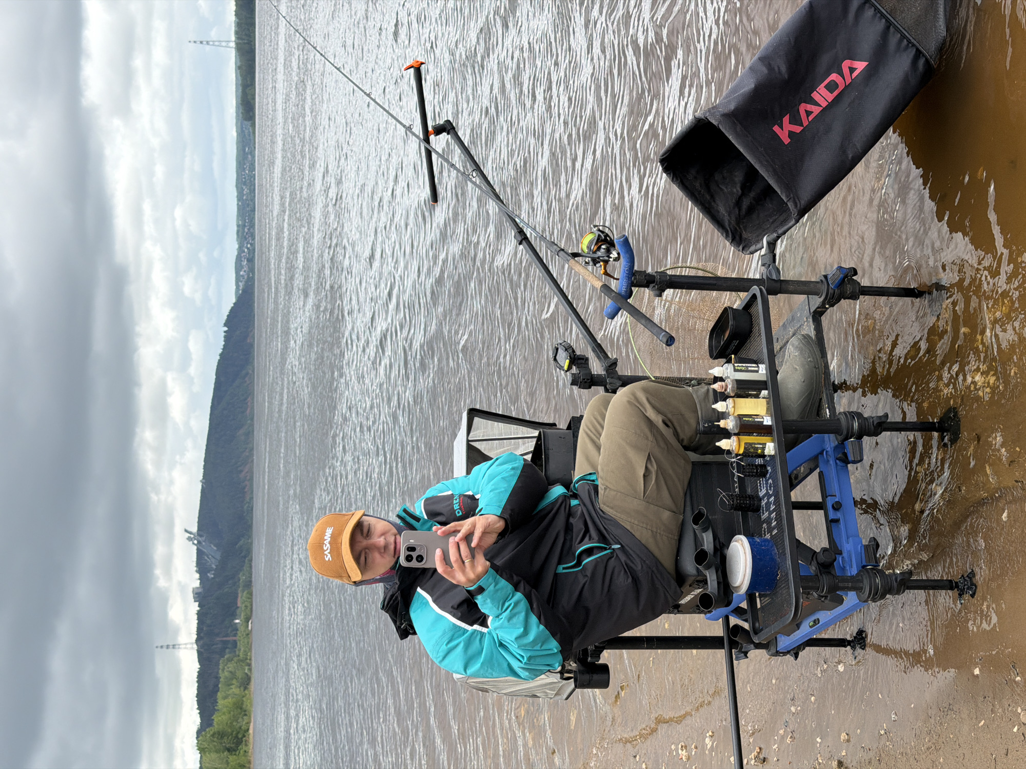 An adult male is seated on a multi-legged blue and black fishing platform, partially submerged in the murky, choppy water of a large river or reservoir. The scene is located in Chaikovsky, Russia, with forested hills visible on the distant shore. On the left hill, a tall transmission tower and a large, angular industrial structure (possibly part of the Votkinsk HPP) are visible. Further right, a smaller tower and a settlement line the horizon.
The man wears an orange baseball cap with "SASAME" in white text, a black and teal waterproof jacket with "DREN" partially visible, olive green trousers, and dark boots. He holds a grey smartphone with both hands, looking at the screen. A ring is visible on his left ring finger.
A fishing rod with a yellow-lined spinning reel and a blue "PRESTON" branded grip is secured in two rod rests, extending over the water. A black mesh side tray attached to the platform holds a white container with a blue lid, several small black fishing weights/feeders, and five small bottles of liquid fishing additives. A black fabric bag with "KAIDA" in red lettering is partially submerged in the water to the right of the platform. The immediate shoreline is wet, muddy, and pebbly. The sky is overcast with grey clouds.