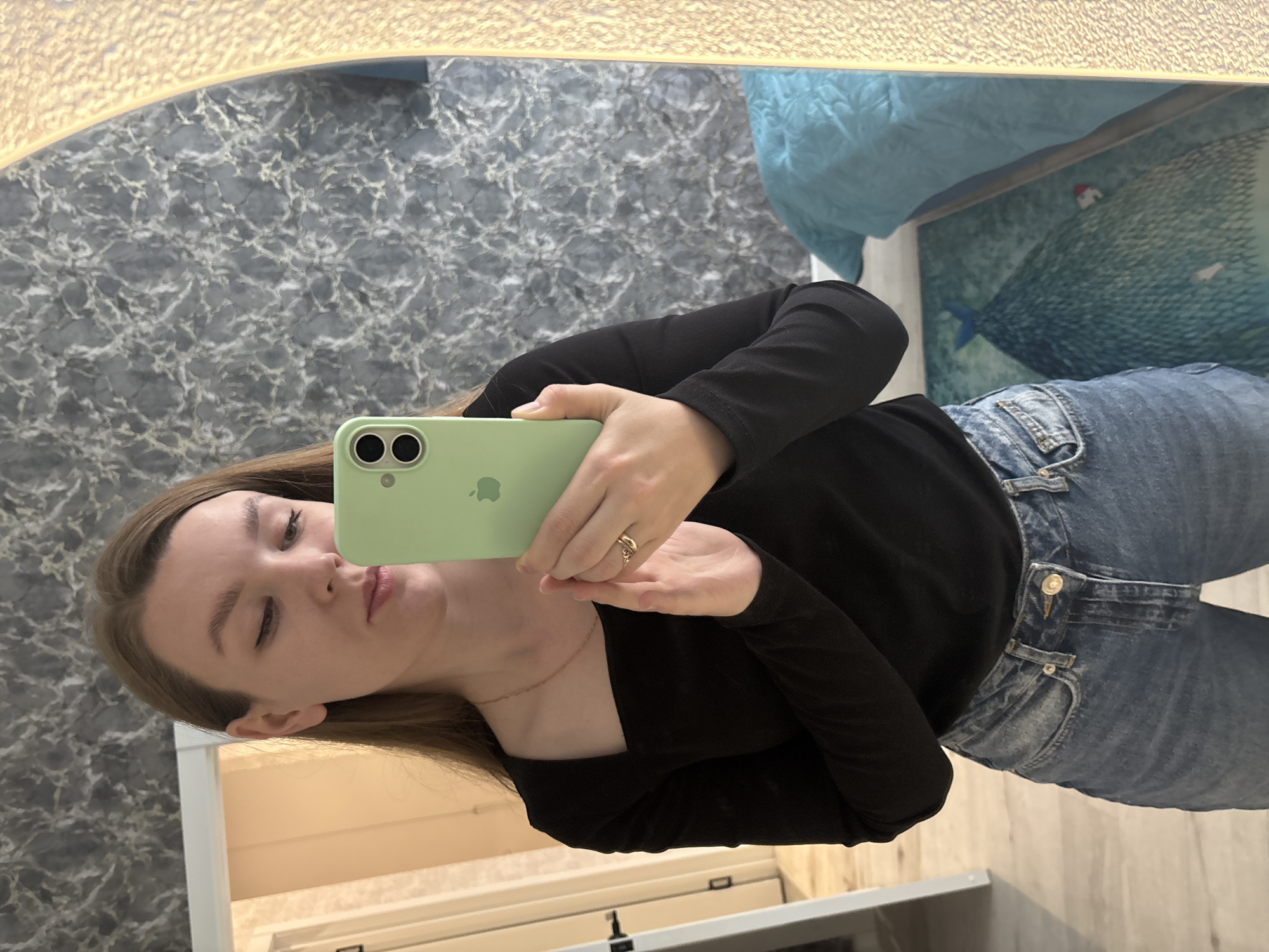 A woman stands indoors, seemingly in a bathroom or a similar domestic space, reflected in a large mirror. She holds up a light green iPhone. She is wearing a black long-sleeved shirt and blue jeans. The background features marble-patterned wallpaper and a light-colored vanity. There's a blue object, possibly a bed covering, partially visible in the upper right. There are no signs in the image that identify the city of Izhevsk, Russia.