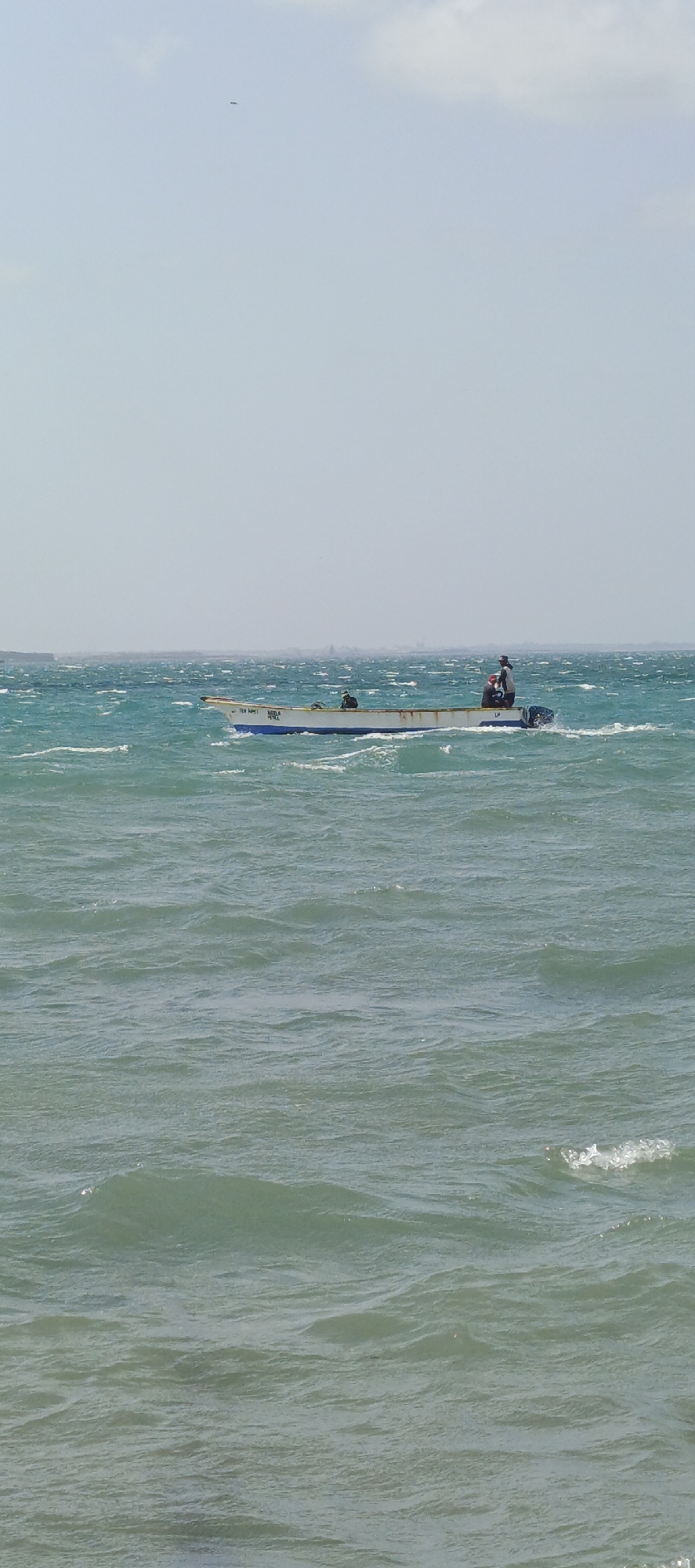 The image depicts a small, open motorboat with three people aboard, navigating choppy waters. The boat is white with a blue trim and the name "TEN NAMI" is visible on its side. The sea is turquoise with visible waves, suggesting a windy day. The sky is light blue with a few faint clouds. The photo appears to be taken from the shore, with the horizon visible in the background. This is Villa Marina in Venezuela.