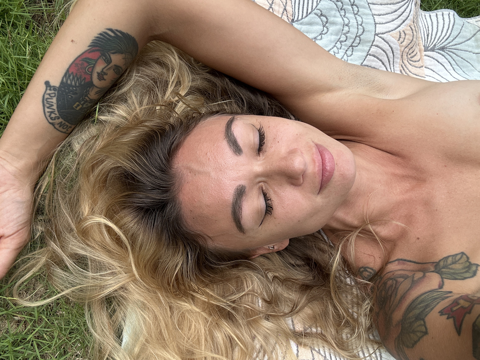 A woman is depicted lying outdoors, appearing to be resting or napping, with her eyes closed and a peaceful expression. She has long, wavy blonde hair with darker roots, fanned out around her head. Her body is lightly tanned, and her arms are casually positioned.
She is lying on a light-colored, patterned blanket or towel, which features geometric and floral designs in shades of white, gray, and peach. Beneath the blanket, lush green grass is visible, indicating an outdoor setting.
Notable tattoos are visible on both arms. On her right arm (viewer's left), there is a prominent tattoo of a punk-style woman's head with spiky black hair and a red background. Below the head, a banner displays the text "PUNKS NOT", with the subsequent word partially obscured but likely "DEAD". On her left arm (viewer's right), part of a larger tattoo featuring green leaves and other design elements is visible.
The natural, bright lighting suggests it is daytime. The overall scene conveys a sense of tranquility and relaxation, set outdoors in Mũi Né, Vietnam.