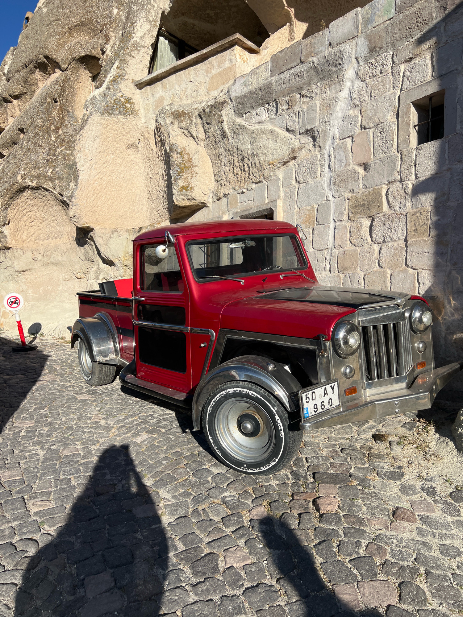 A red vintage pickup truck is parked on a cobblestone street in Göreme, Turkey, with ancient rock formations and stone buildings in the background. The truck has a chrome grille, fenders, and bumper. A license plate on the front reads "50 AY 960" with a Turkish flag. To the left of the truck is a red and white sign with a no-parking symbol. The sun casts a long shadow of the photographer onto the cobblestones. The rock formations appear to be part of the unique landscape of Cappadocia, characterized by fairy chimneys and cave dwellings. The stone building to the right has a small window and textured walls.