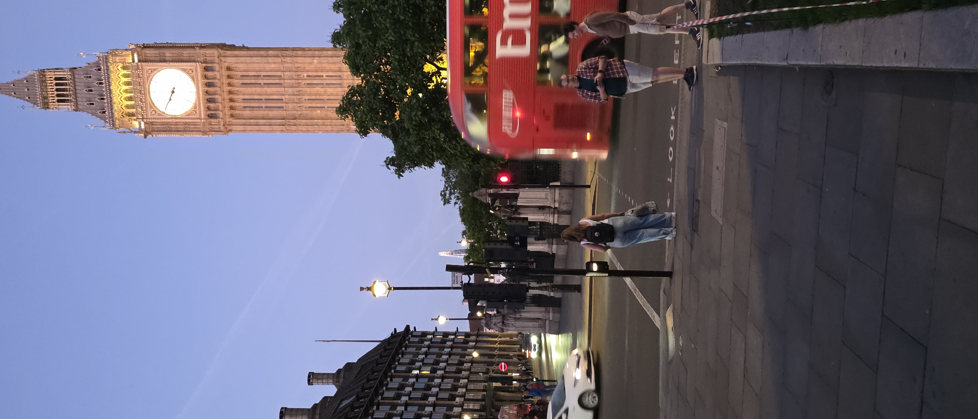 The image depicts a street scene in London, United Kingdom, featuring the iconic Elizabeth Tower (often referred to as Big Ben) prominently in the background. The sky is a clear twilight blue, suggesting it is early evening. The Elizabeth Tower, illuminated from within, stands tall on the right side of the frame.
In the mid-ground, a red double-decker bus is in motion, blurred by its movement. On the sidewalk to the right of the bus, two men are standing. One man, wearing a plaid shirt and shorts, is looking at his phone. Next to him, another man in a brown sweater and shorts is walking.
On the left side of the street, a white car is also in motion, blurred due to its speed. A woman with a backpack and carrying shopping bags stands on the corner, facing away from the camera, waiting to cross the street. Behind her, traffic lights are visible. Further to the left, a large building with numerous windows lines the street. Streetlights with warm yellow illumination are present along the road, casting a soft glow. In the far distance, a modern skyscraper with a pointed top is visible on the horizon. The street is paved with stone slabs. Red and white striped tape runs along the edge of the sidewalk on the right side of the image.