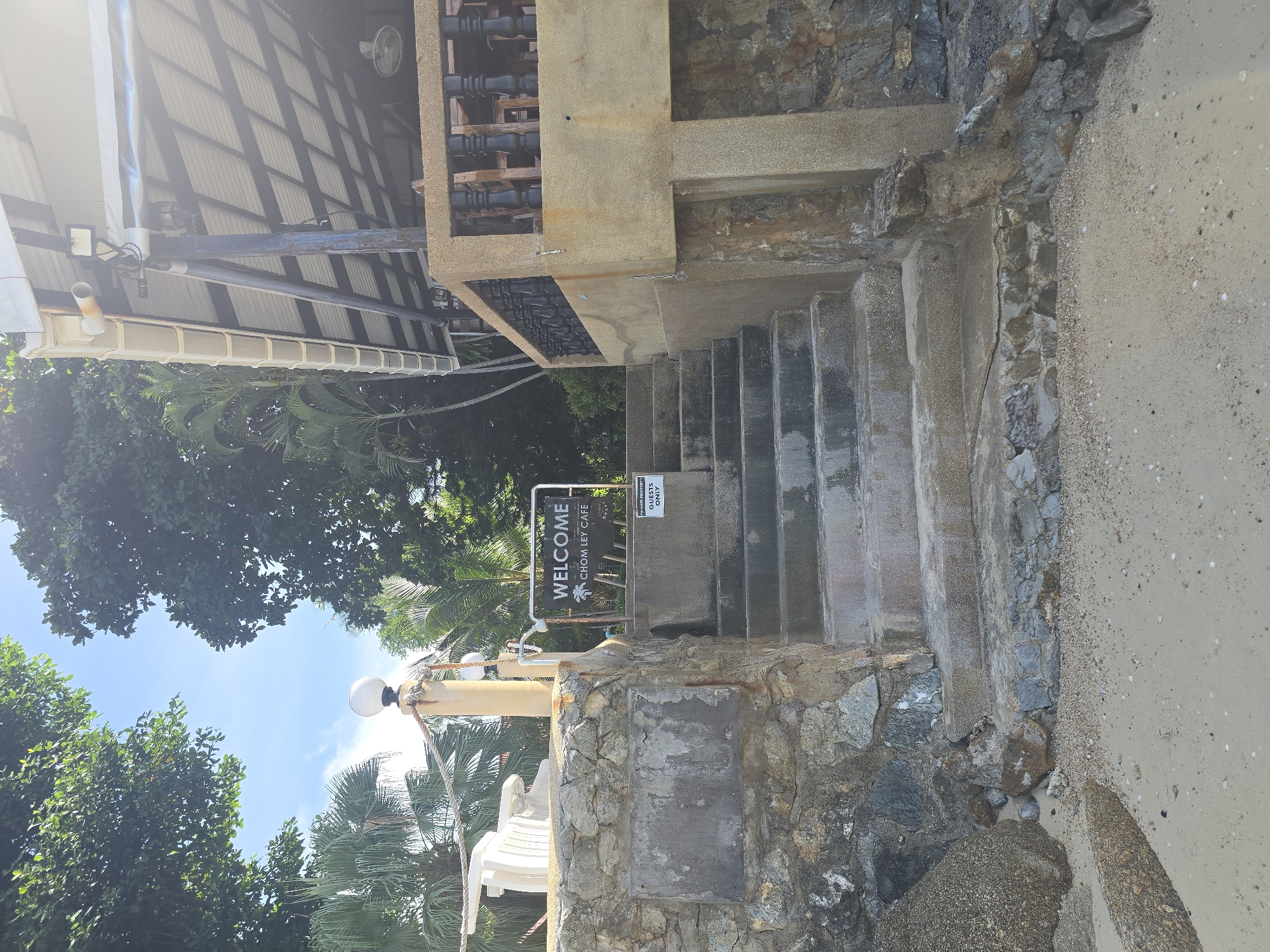 The image shows an outdoor scene at Chom Ley Cafe in Baan Chaweng Noi, Thailand. A stone staircase leads up to a building with a covered outdoor seating area. A "WELCOME CHOM LEY CAFE" sign is visible at the top of the stairs, along with a smaller sign that reads "PRIVATE PROPERTY GUESTS ONLY". Trees and palm trees are visible in the background. The area appears to be on a beach, with sand and rocks in the foreground. There are no people visible in the image. The sky is bright and sunny.