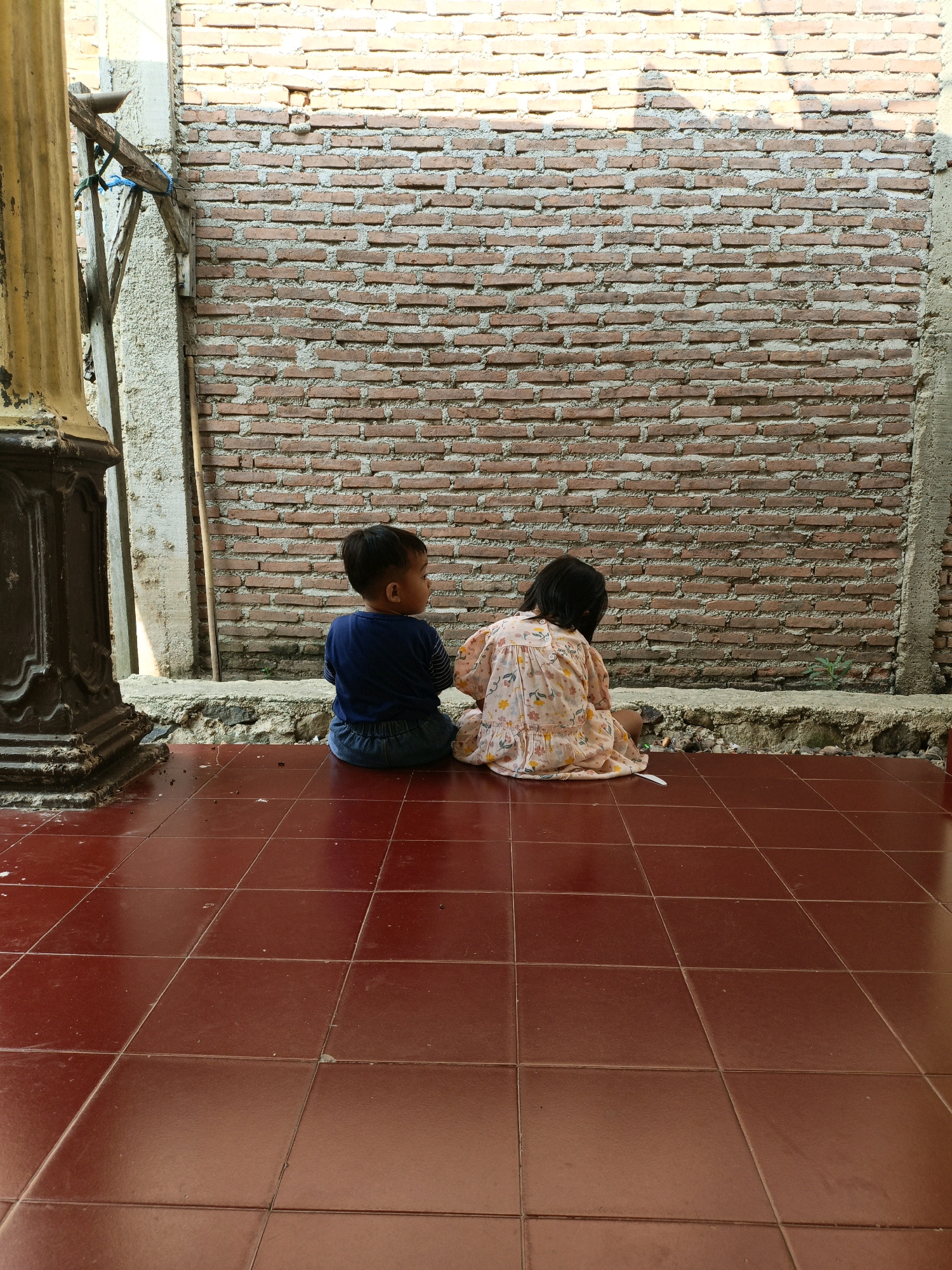 In the city of Bunut, Indonesia, a young boy and girl are seated on a tiled floor, facing away from the camera. The boy, wearing a blue shirt and denim shorts, is positioned to the left. To his right, the girl is dressed in a light-colored, patterned dress. They are situated in front of a brick wall, partially obscured by structural elements and shadows. The foreground is dominated by a red tiled floor.