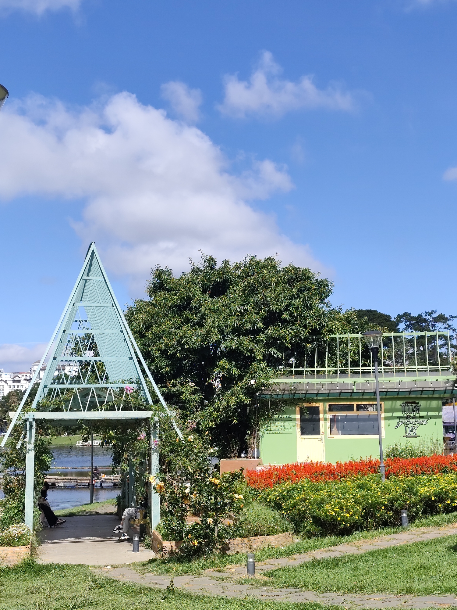 A sunlit, vertical medium shot captures a picturesque scene in Da Lat, Vietnam. A blue sky, dotted with fluffy white clouds, forms the backdrop. In the foreground, a green metal structure with a triangular top stands over a small pathway leading towards a body of water. People are visible sitting or relaxing within and around the structure. Lush greenery, including flowering plants, surrounds the pathway. Adjacent to the pathway is a light-green building with a sign that reads "Today is a good day." A vibrant array of red and yellow flowers lines the front of the building. The scene is framed by grass and paved walkways. The environment suggests a park-like or garden setting, with the city buildings visible in the distance.