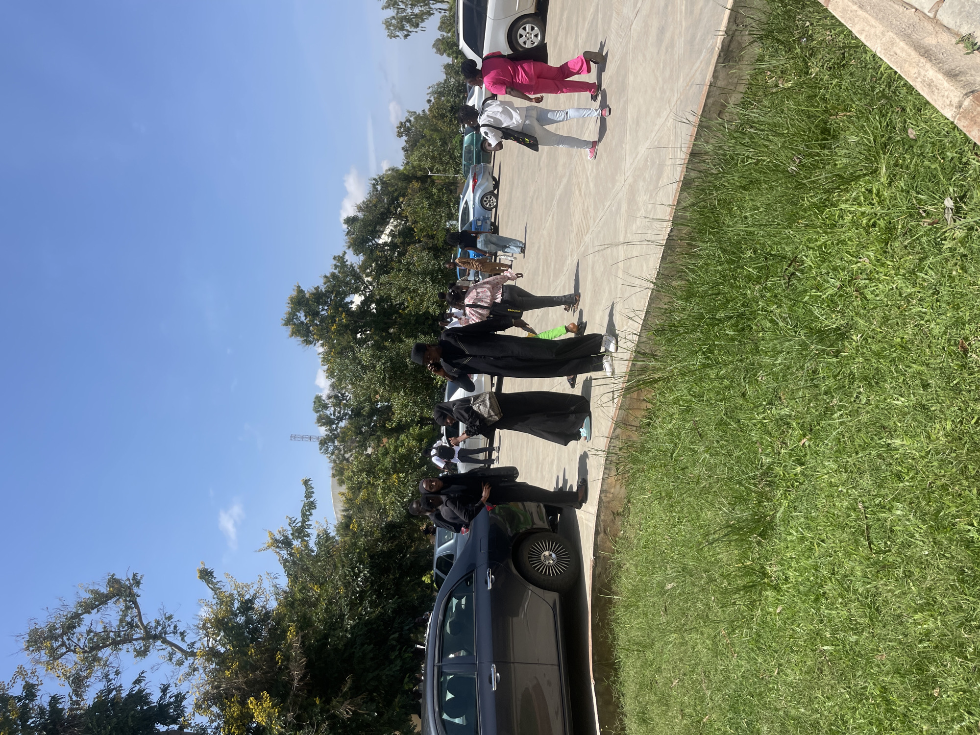 In a paved outdoor area in Rafin Par, Nigeria, several individuals are visible near parked cars under a clear blue sky. The scene appears to be a parking lot or courtyard, with a dark sedan on the left and other vehicles further back. A group of people, some wearing black attire and headscarves, are walking and standing. One woman in a pink outfit is walking away from the camera. Trees with green foliage and some yellow blossoms are present in the background, along with a building and a tower structure. The ground is concrete, bordered by a strip of green grass in the foreground.
