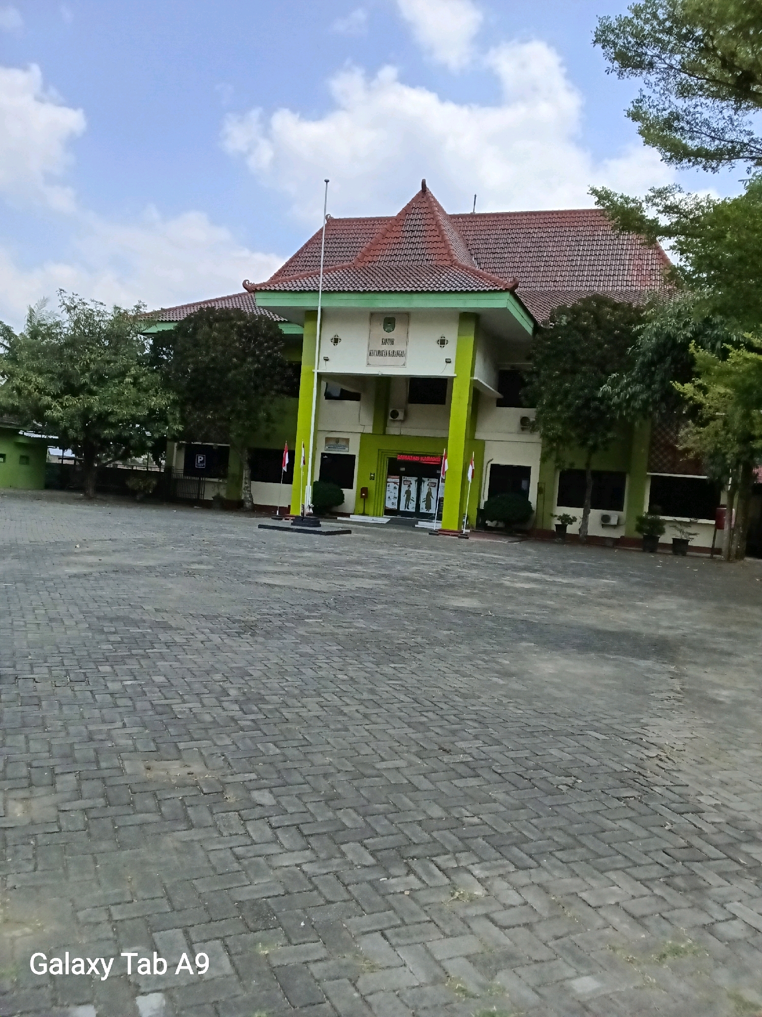 The image shows a two-story building with a red tiled roof, characteristic of Indonesian architecture, in the city of Karangan, Indonesia. The building has a green facade with white trim and prominent lime green pillars supporting an overhang. The entrance is flanked by two flagpoles, one displaying the Indonesian flag. Above the entrance, a sign reads "KANTOR KECAMATAN KARANGANGA". The building is situated in an open, paved area, likely a public square or parking lot, with brick-like paving stones. Trees surround the building, and the sky is partly cloudy. The foreground of the image displays the text "Galaxy Tab A9". There are no people visible in the image. The scene depicts a government administrative building on a clear day.